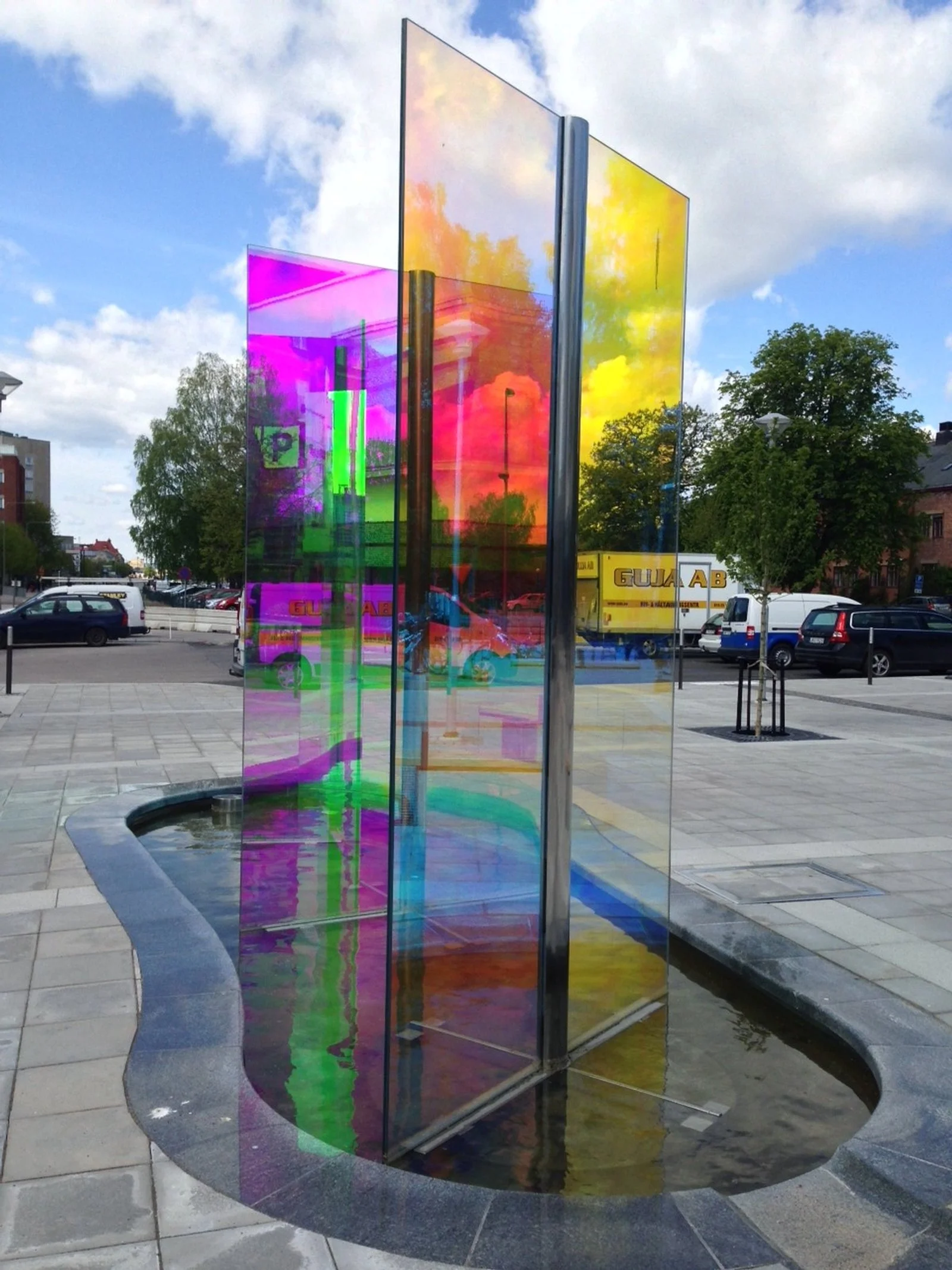 Colorful reflective glass sculpture with multiple panels, reflecting the sky and surroundings, in an urban plaza.