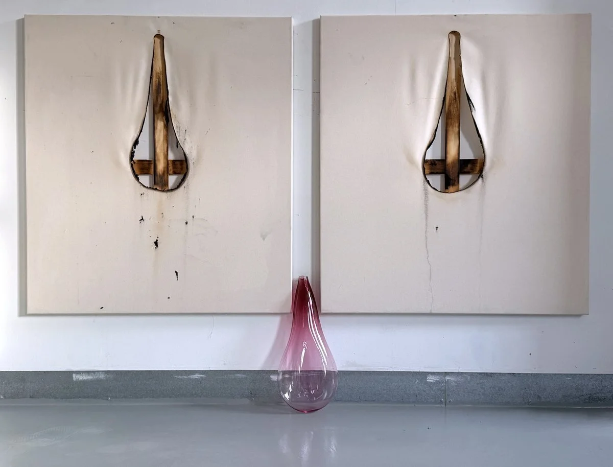 Two white canvas panels with burnt wooden teardrop-shaped cutouts, leaning against a white wall, with a pink glass vase on the floor between them.