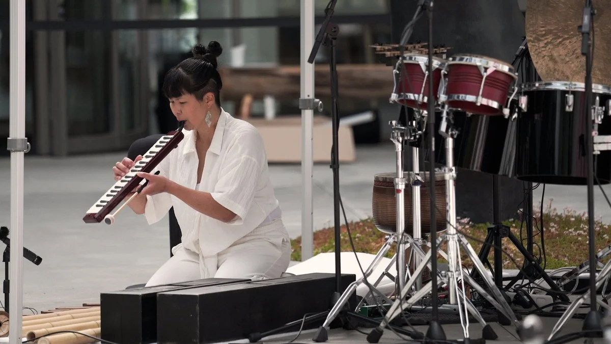 A woman in white clothes playing a melodica during an outdoor musical performance, with drums and percussion instruments visible nearby.