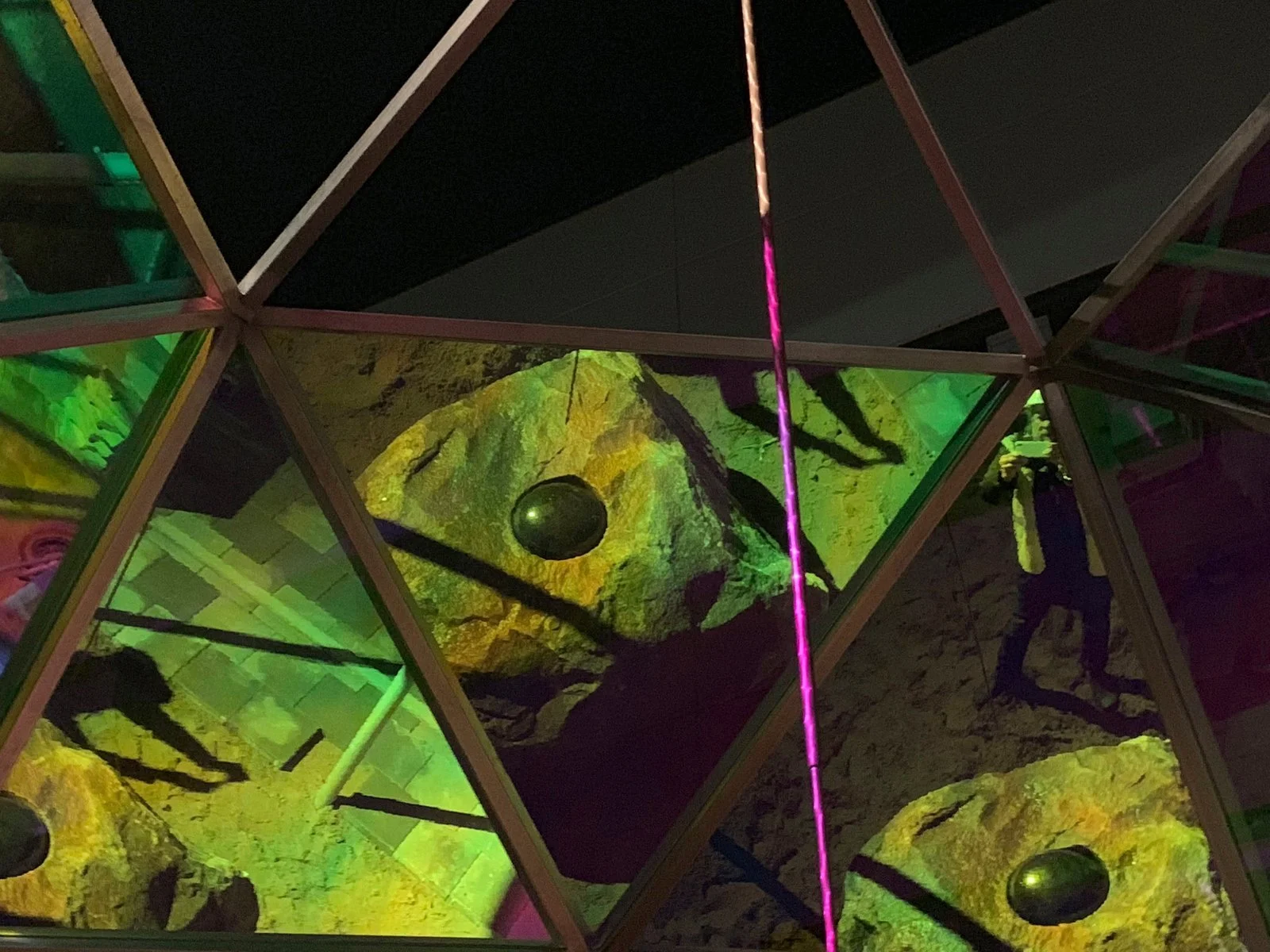 Interior view of a climbing gym with rocks positioned underneath a glass ceiling, illuminated by colorful lighting, with a person climbing and shadows cast on the rocks.