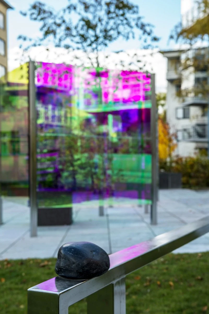 A black rock placed on a silver metallic railing with a colorful reflective glass installation and trees in the background.