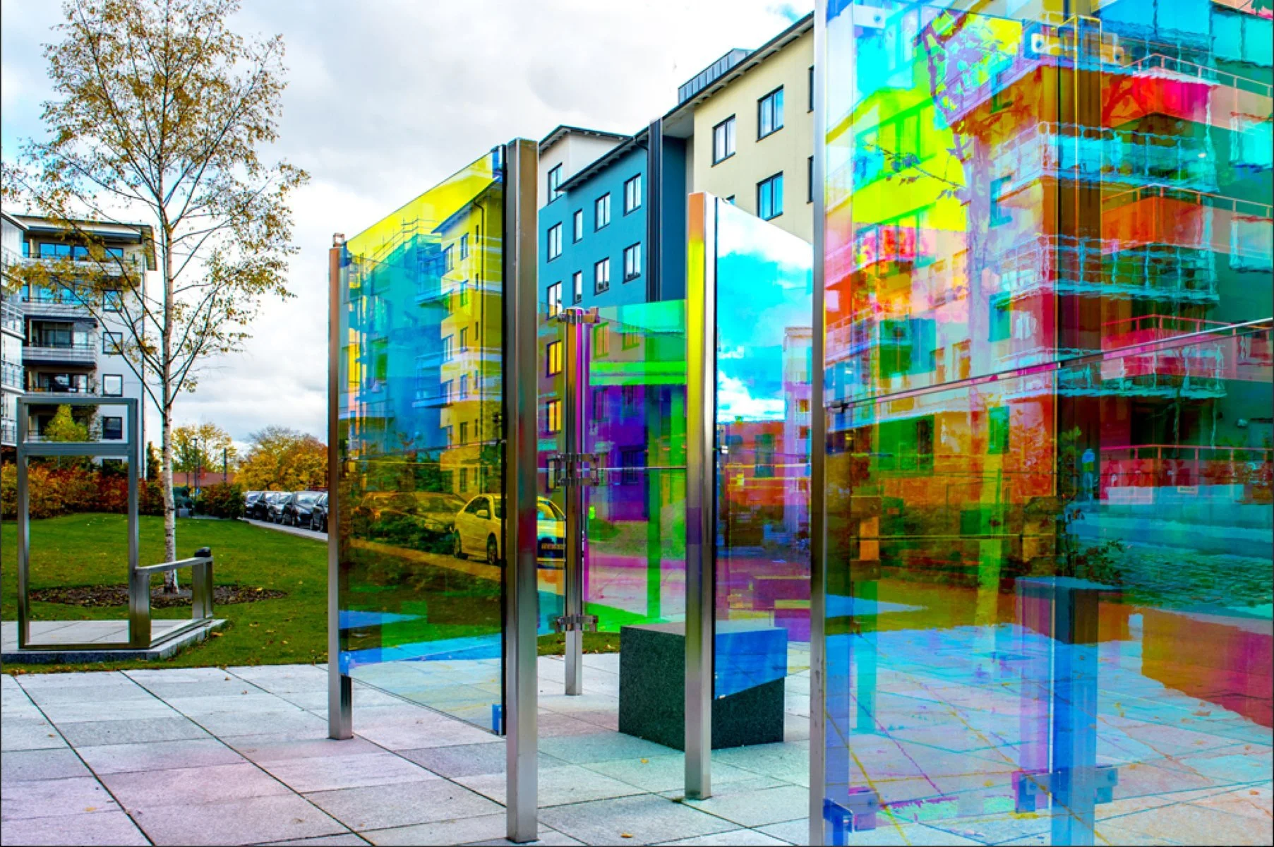 Colorful glass art installation on city sidewalk with apartment buildings, trees, and parked cars in the background.