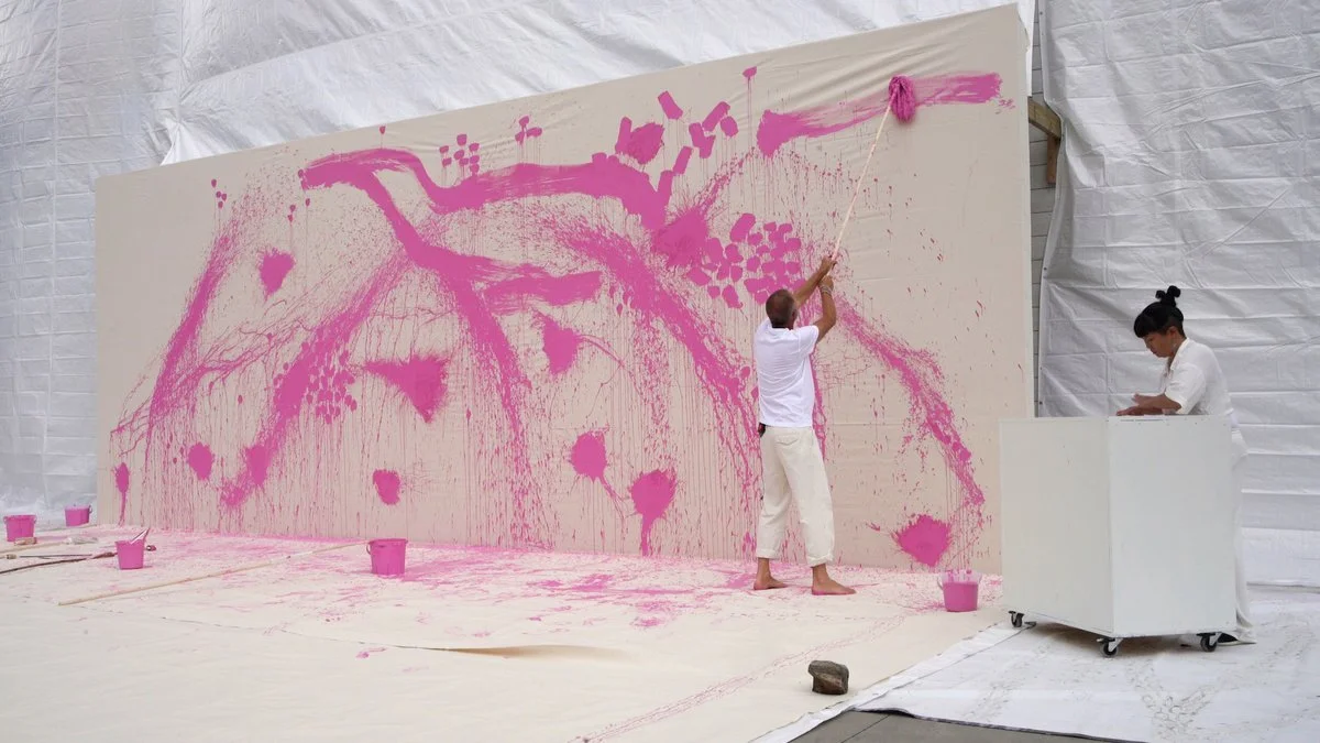 Two artists are creating a large pink abstract mural on a beige wall. One artist is using a mop attached to a stick to apply pink paint, while the other artist is standing nearby, working at a white mobility cart. Several pink paint buckets are on th