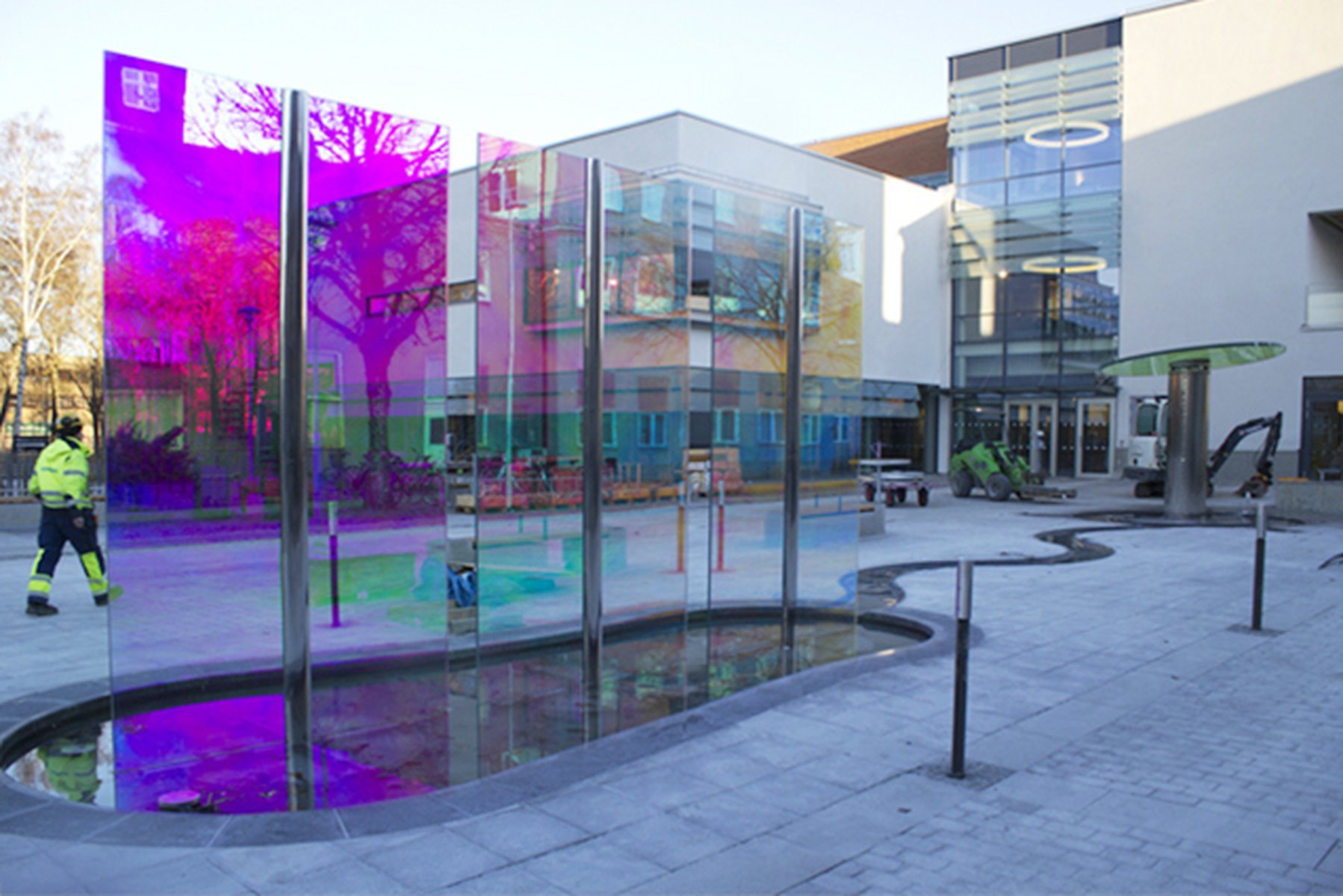 Construction site outside a modern building with glass panels, a worker in a high-visibility vest walking nearby, and equipment including a small vehicle and a hydraulic lift.