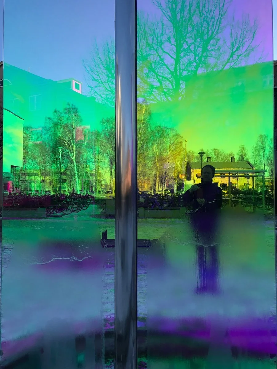 Reflection of a person taking a photo in front of a colorful, iridescent glass surface with trees and buildings in the background.
