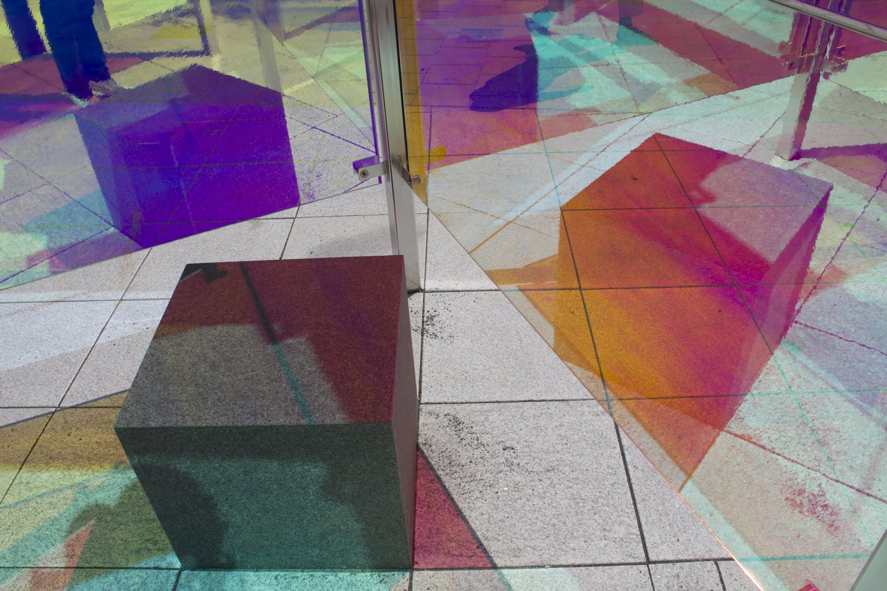 Colorful projection of geometric shapes and patterns on tiled floor, with a dark-colored cube in the foreground and glass doors in the background.