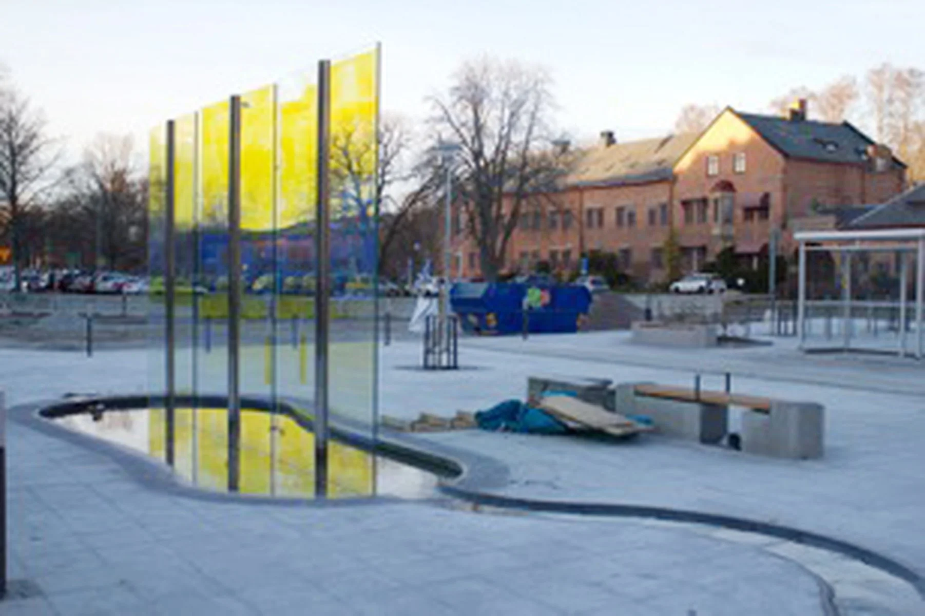An outdoor urban scene with modern art installation, including several tall, yellow, reflective panels, a small water feature, and concrete seating near a trash bin, with buildings and parked cars in the background.