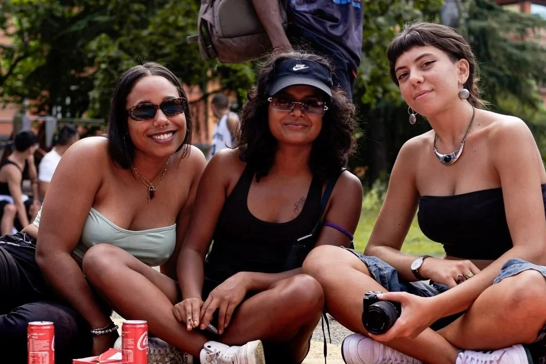 A smiling man wearing sunglasses, a beige cap, and headphones gives a thumbs-up in front of a colorful sign that reads "REGAZ DEI FAVA." He is outdoors with trees in the background.