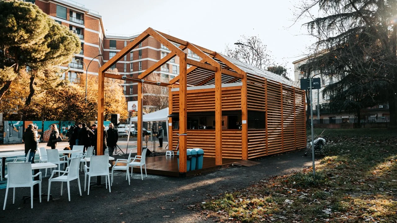 A wooden structure with open framing on top, used as a small event space or pavilion, set up outdoors on a city street with chairs and people around.