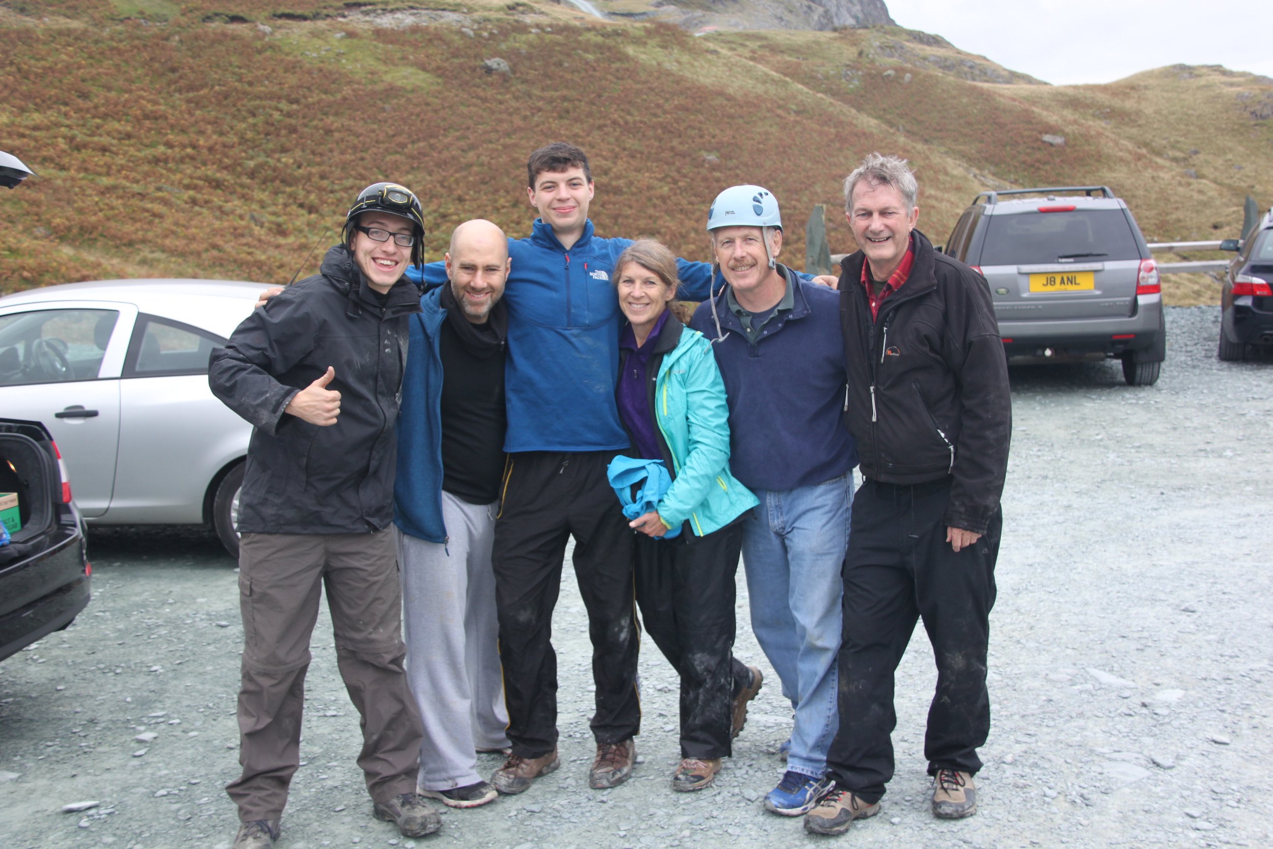 Six people, three men and three women, standing together outdoors in front of parked cars on a gravel surface, smiling and enjoying each other's company, with a hilly, grassy landscape in the background.
