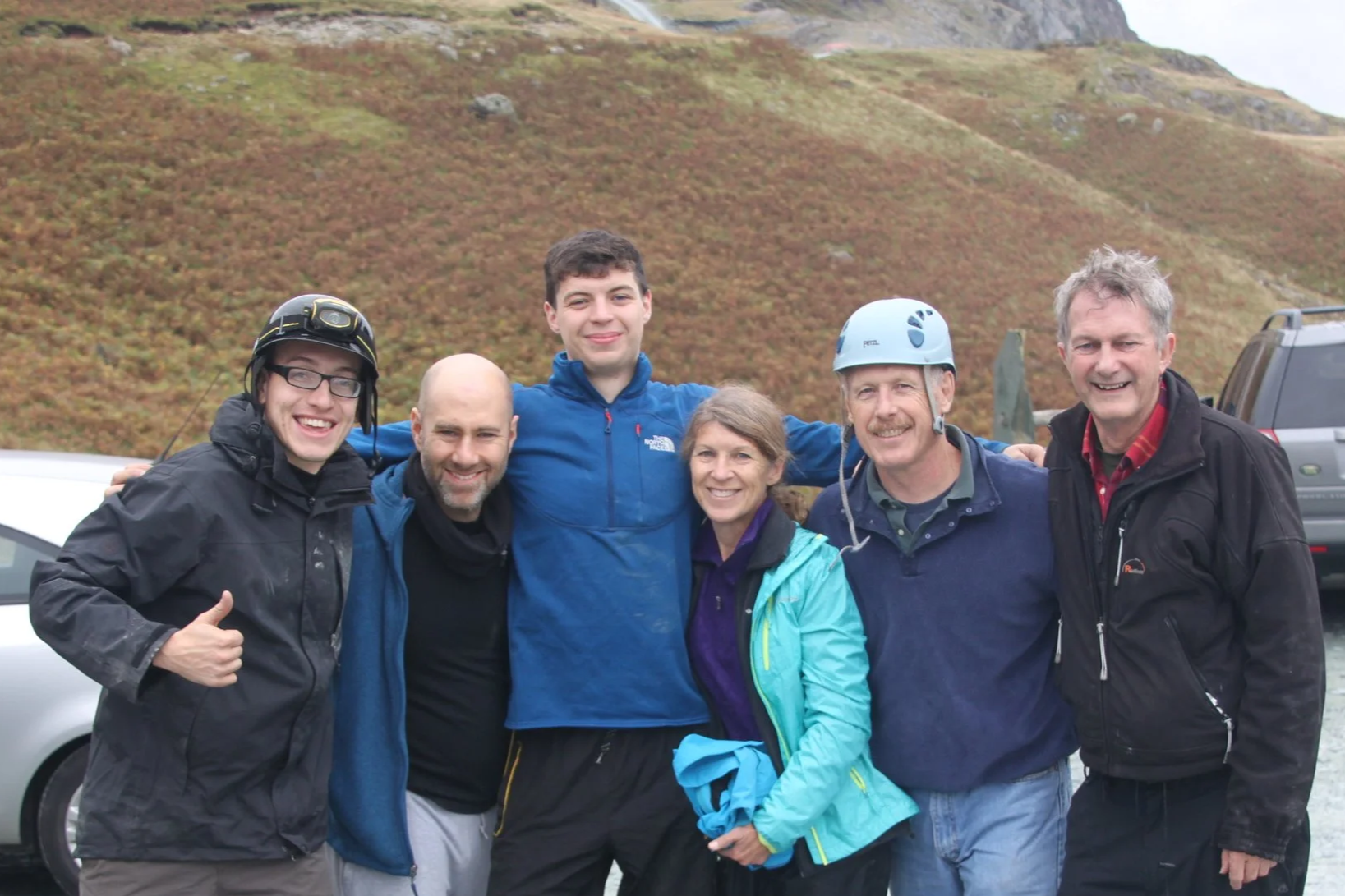 Group of six people smiling outdoors with a hillside in the background, some wearing outdoor and safety gear.