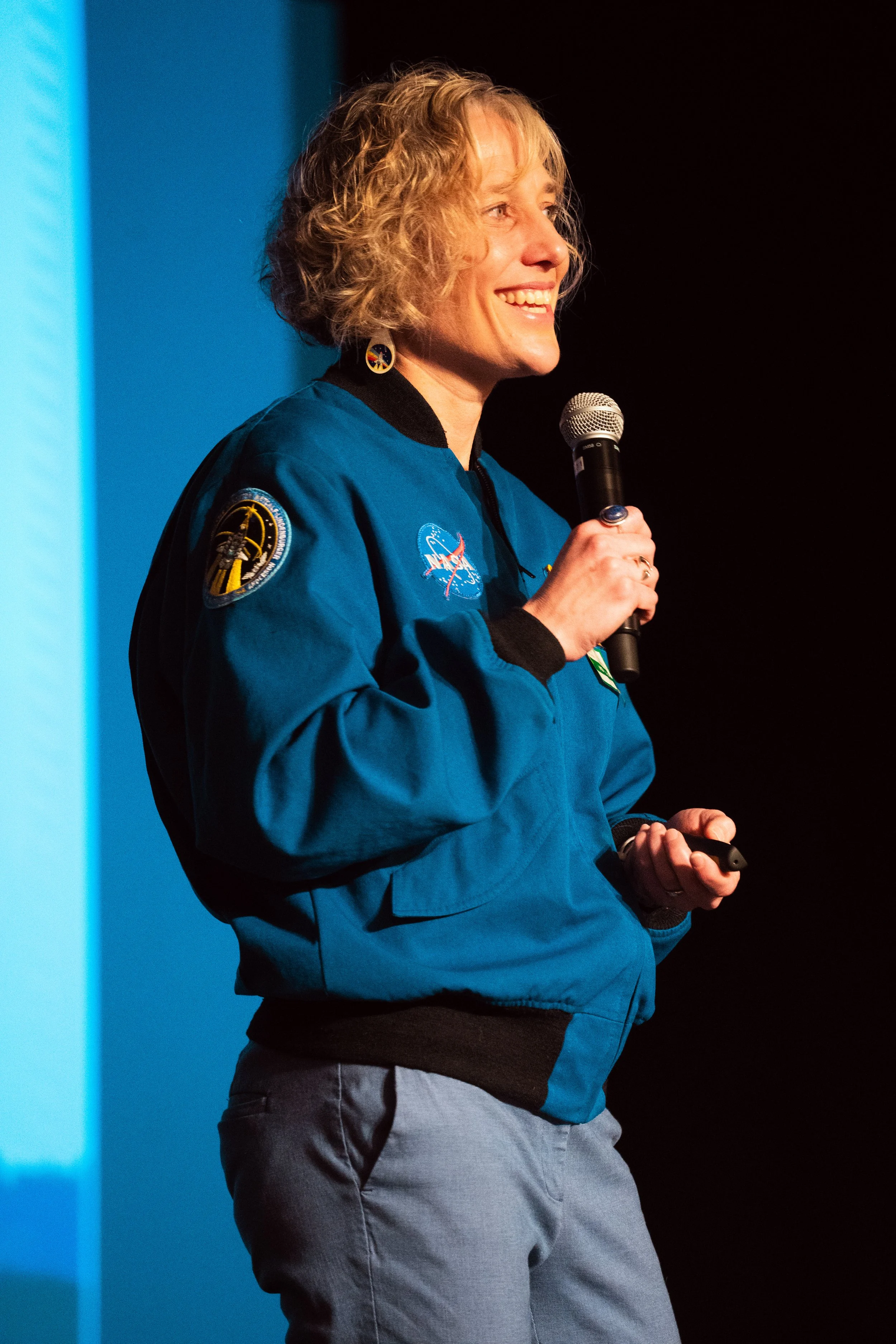 A woman in an astronaut jacket holding a microphone and smiling on stage.
