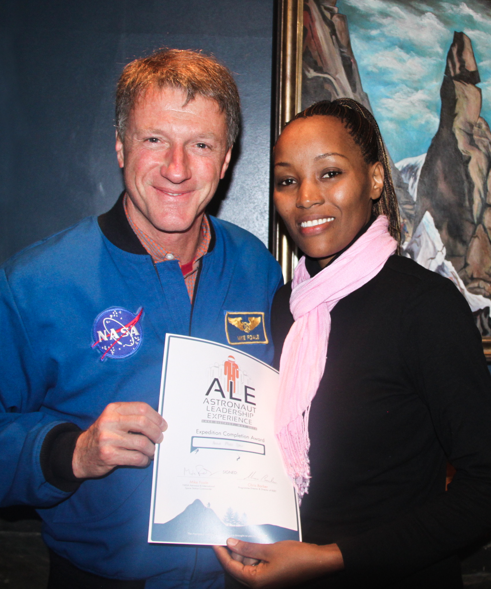 A man in a blue NASA jacket and a woman in a black top and pink scarf holding a certificate for the Astronaut Leadership Experience, with a mountain painting in the background.