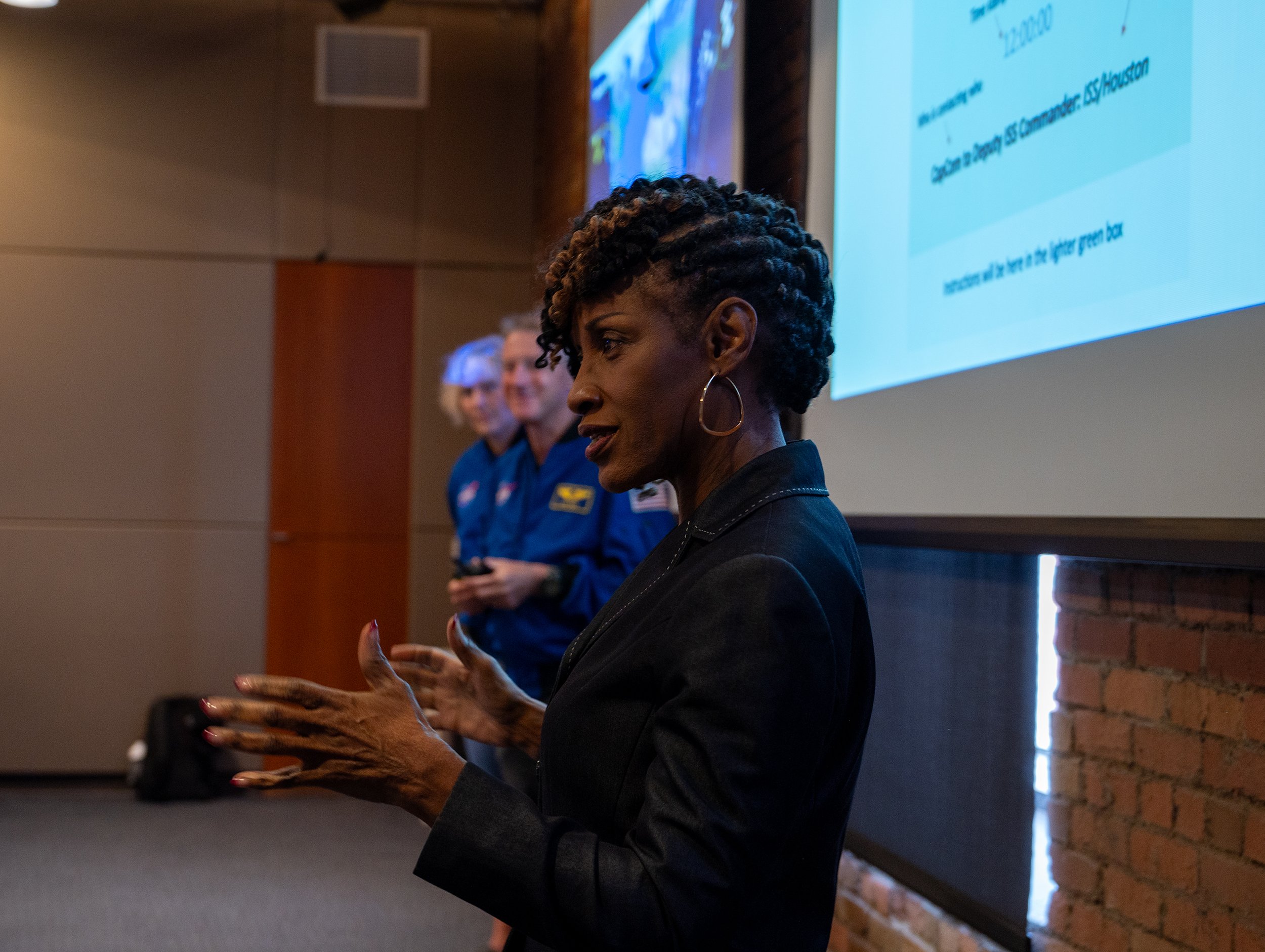 A woman giving a presentation in a conference room, with people in blue NASA jackets standing behind her and a large screen displaying a presentation slide.