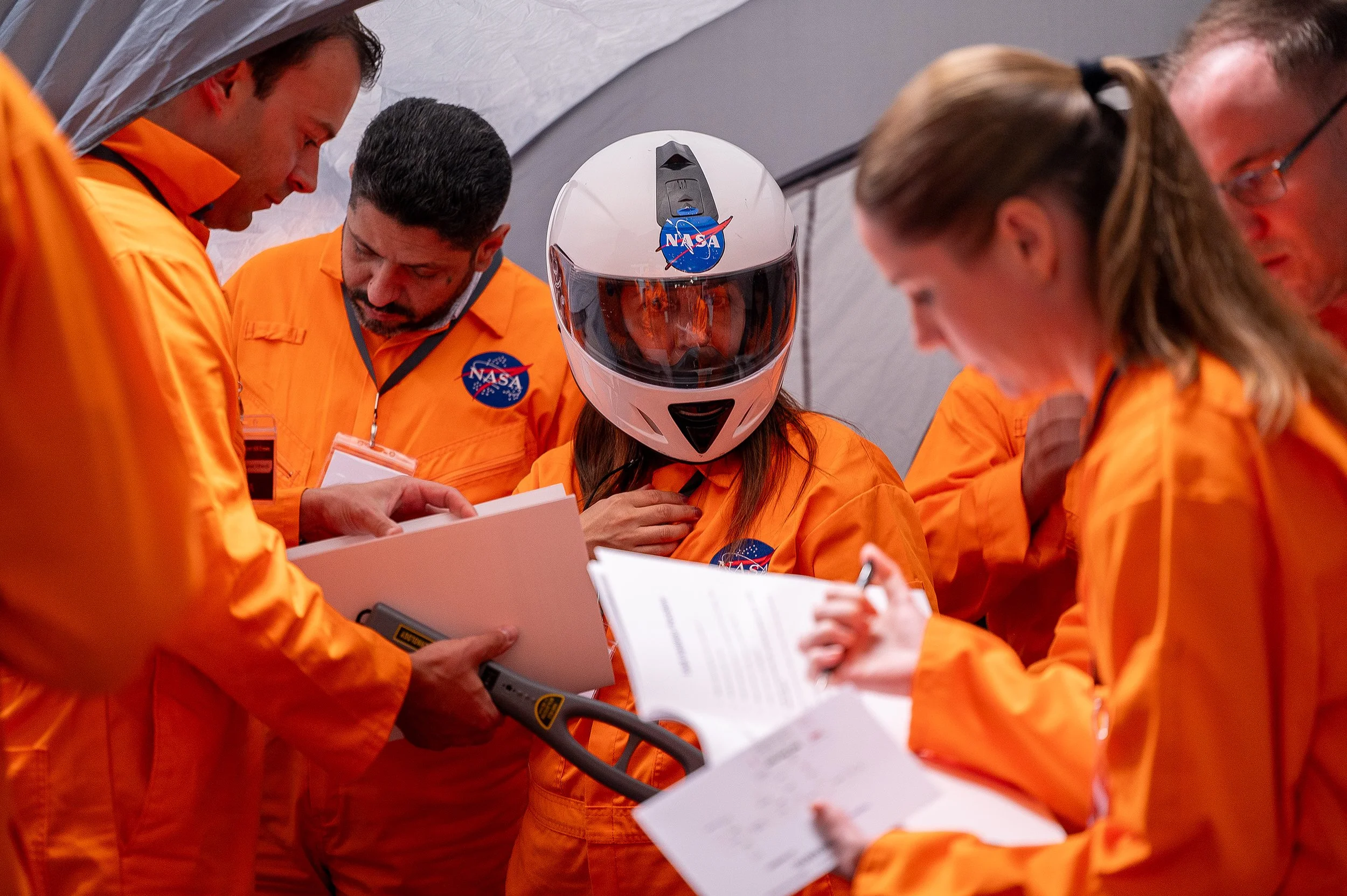 NASA crew members in orange jumpsuits gathered around, discussing plans while one woman wears a white helmet with a visor and NASA logo.