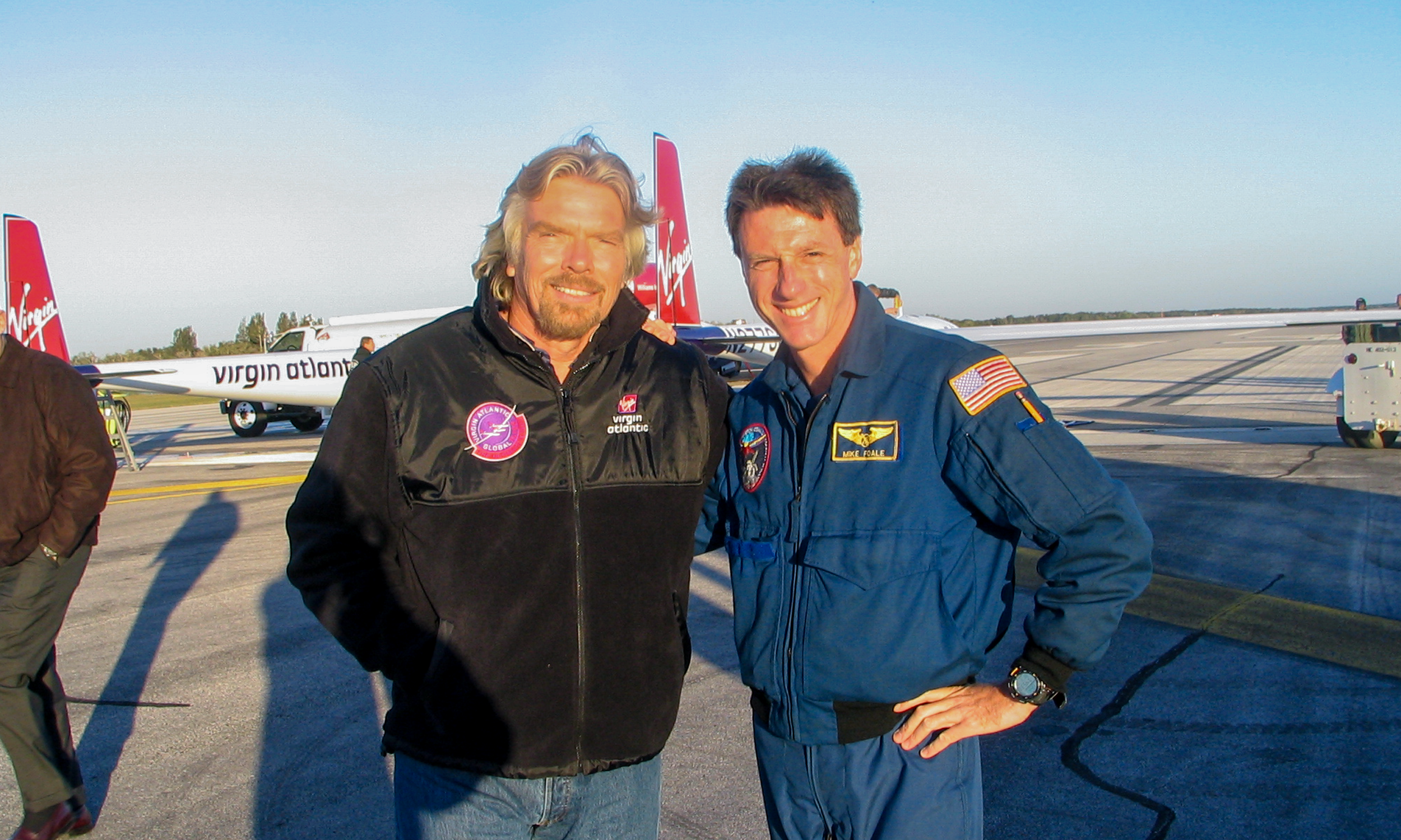 Two men standing together on an airport tarmac, one in a black jacket with Virgin Atlantic logos and the other in a blue pilot uniform with a patch that reads 'Mike Fogle.' Behind them are Virgin Atlantic airplanes and airport ground vehicles.