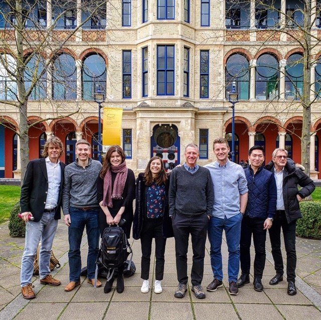 Group of eight diverse people standing in front of a historic brick and stone building with large arched windows, during daytime. They are smiling and dressed casually.