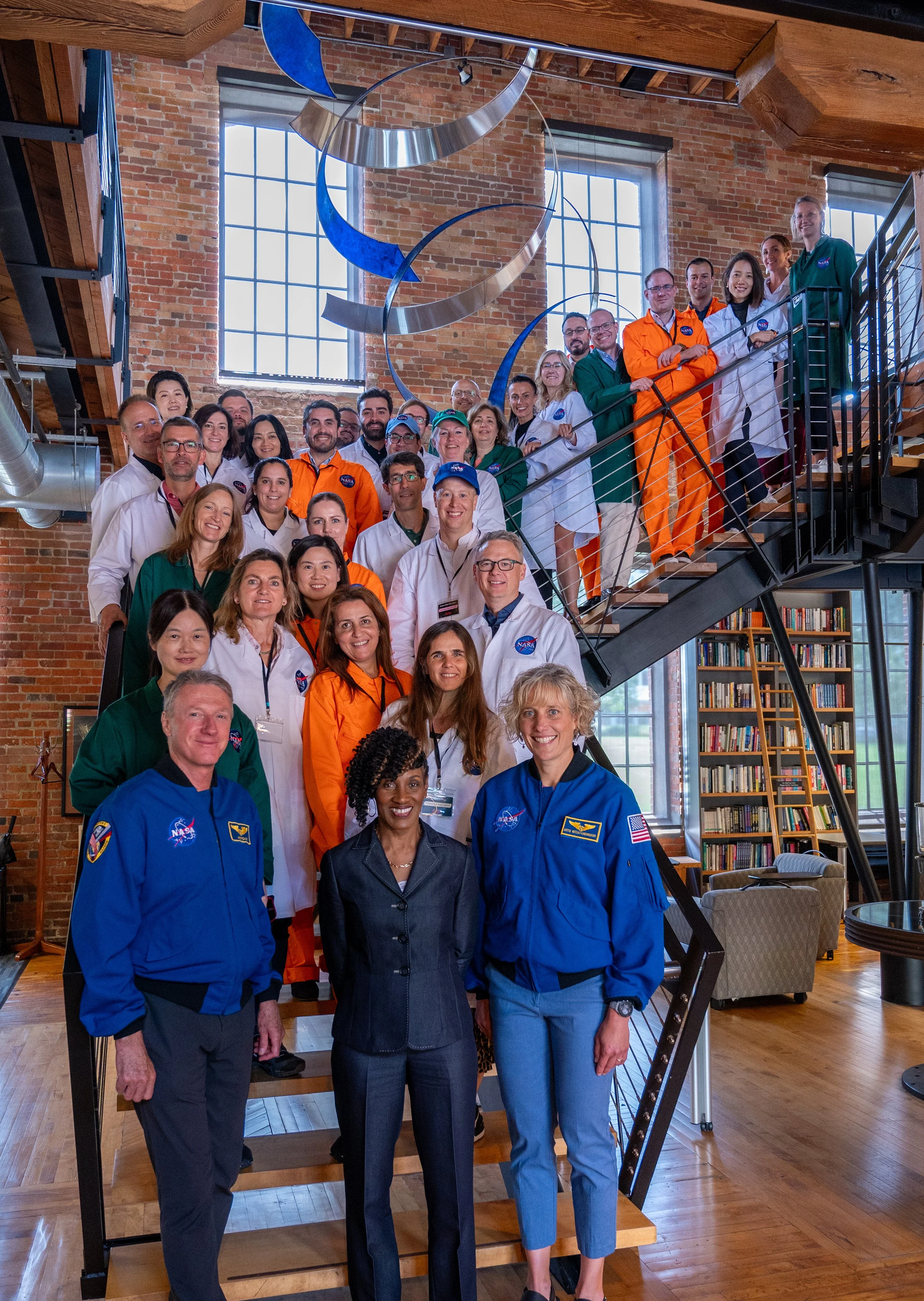Group of NASA astronauts and staff posing on a staircase with brick walls and large windows in the background.
