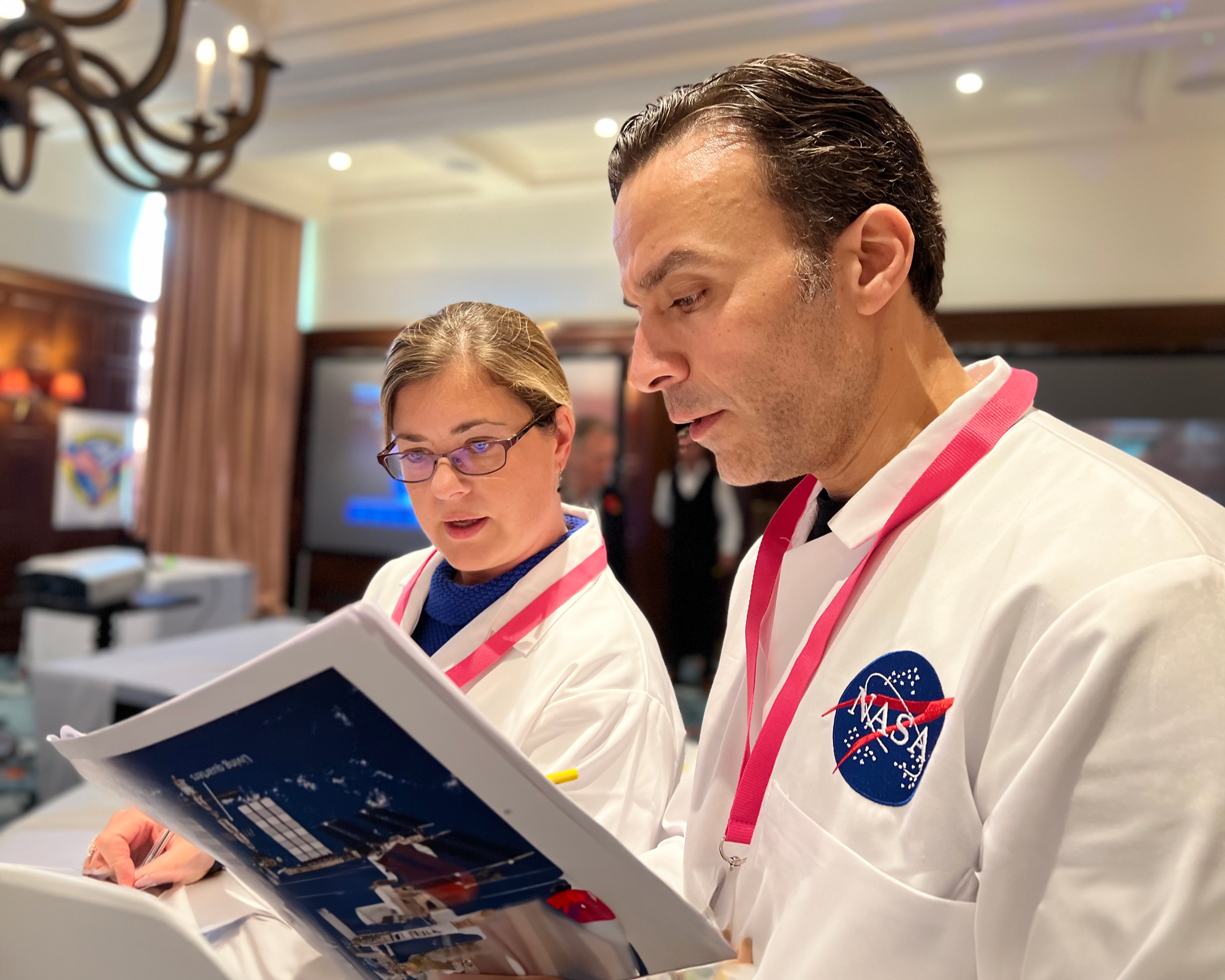 Two astronauts, a woman with glasses and a man, wearing NASA uniforms, reviewing a document inside a room with wood-paneled walls and a chandelier.