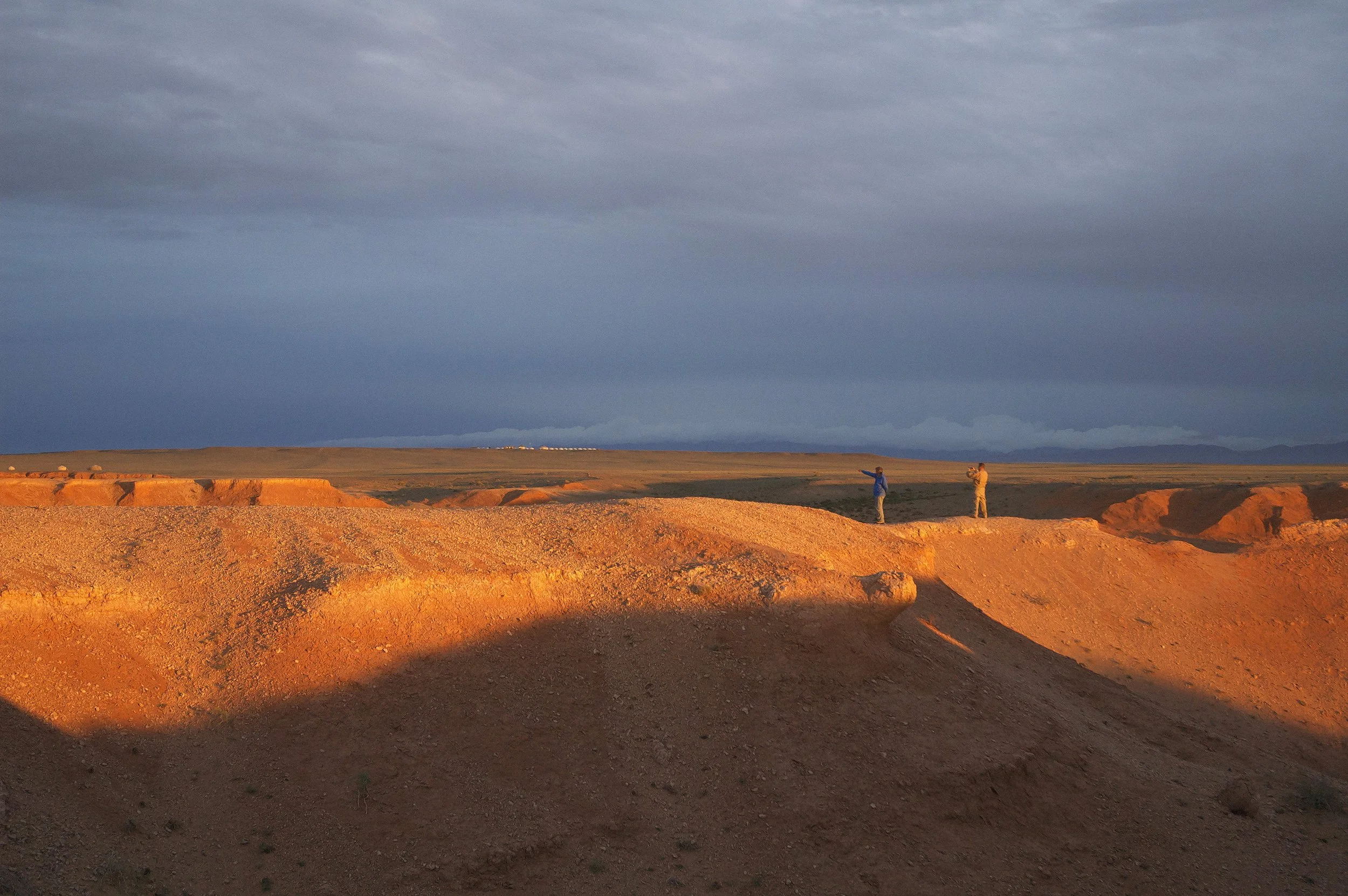 A desert landscape at sunset with two people standing on rocky formations, one pointing toward the horizon under a dark, cloudy sky.