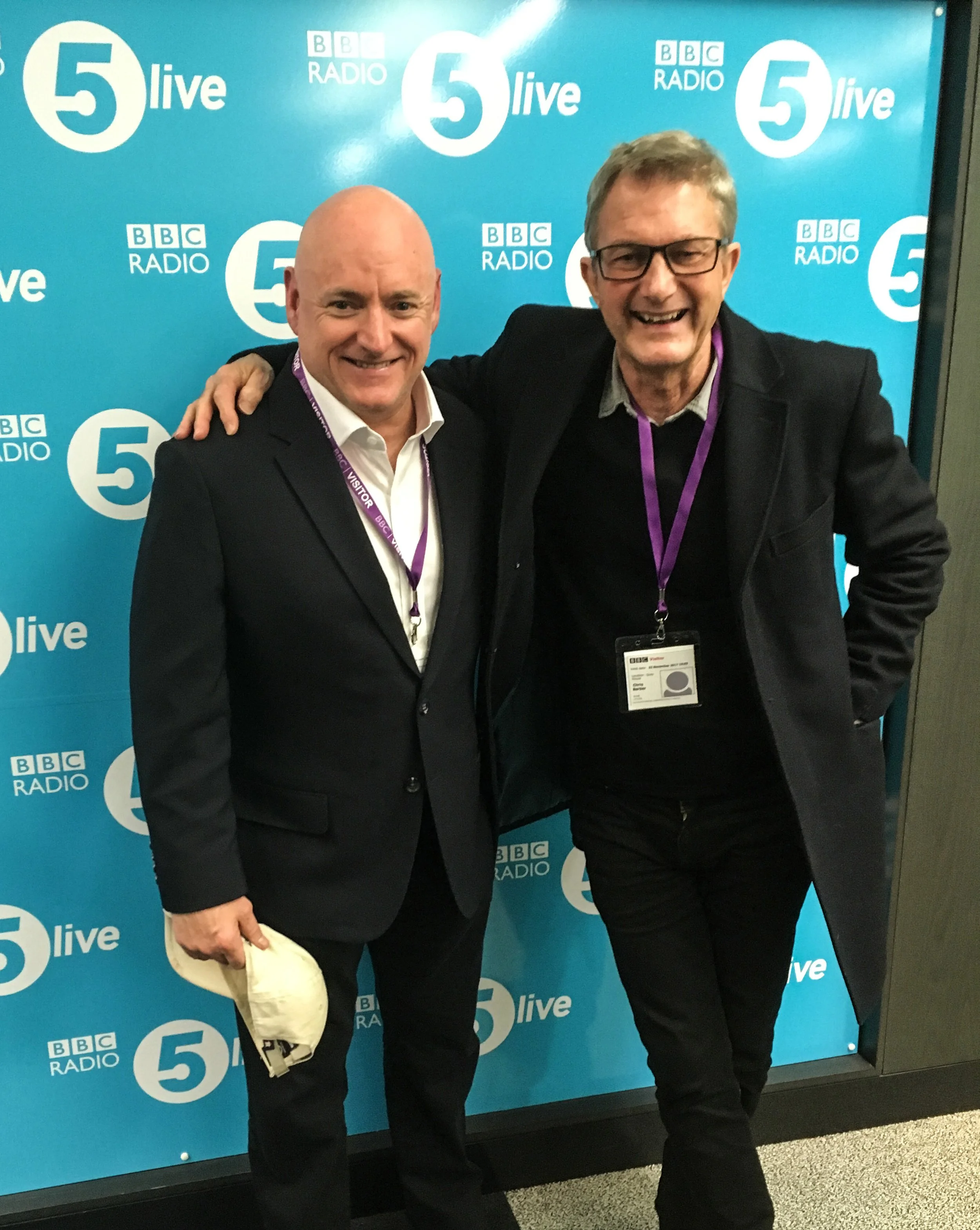 Two men smiling and posing with an arm around each other in front of a BBC Radio 5 live backdrop at a radio station.