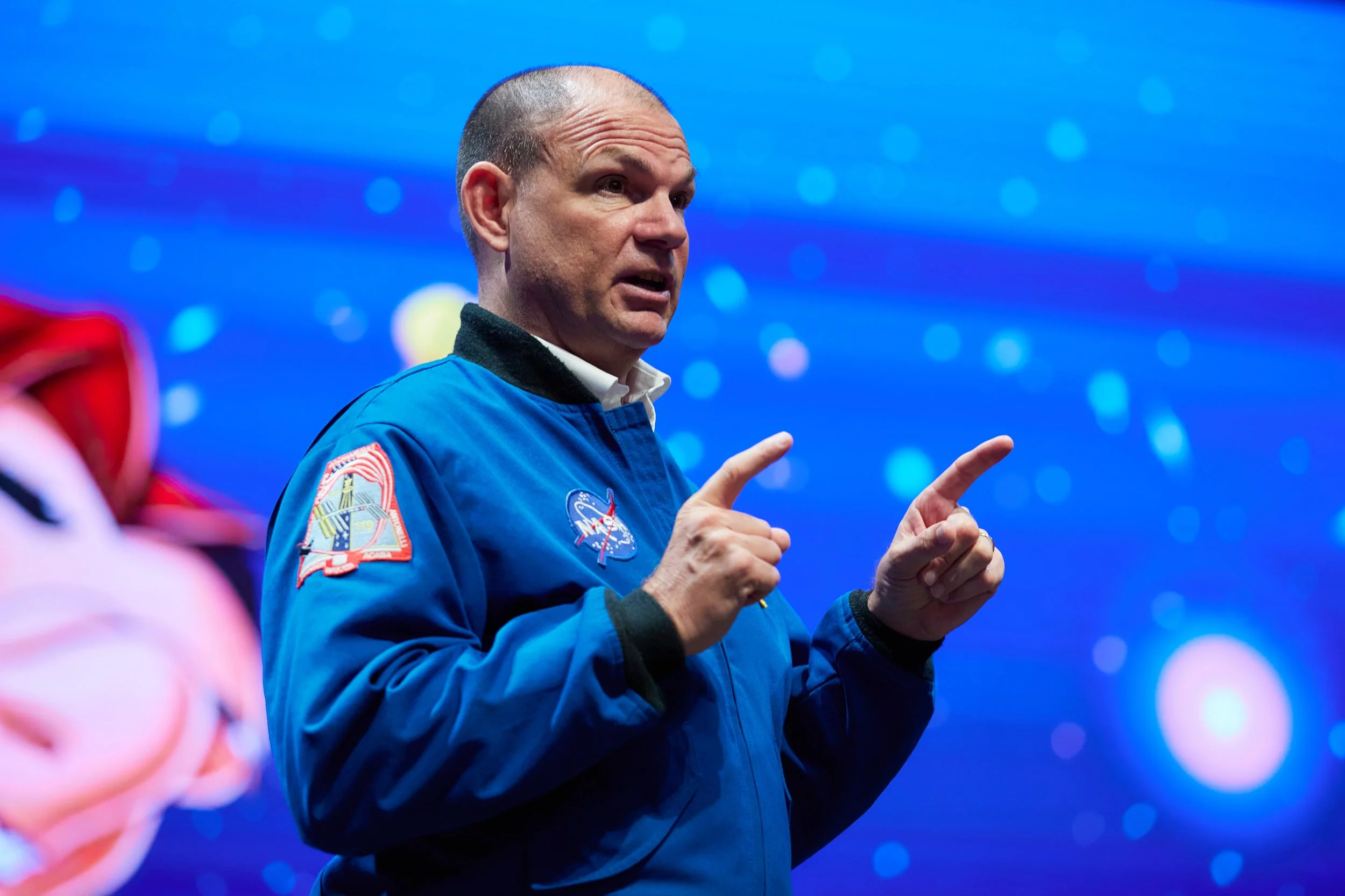 A man in a blue NASA jacket is gesturing with both hands during a presentation or discussion, with a blue galaxy or space-themed background.