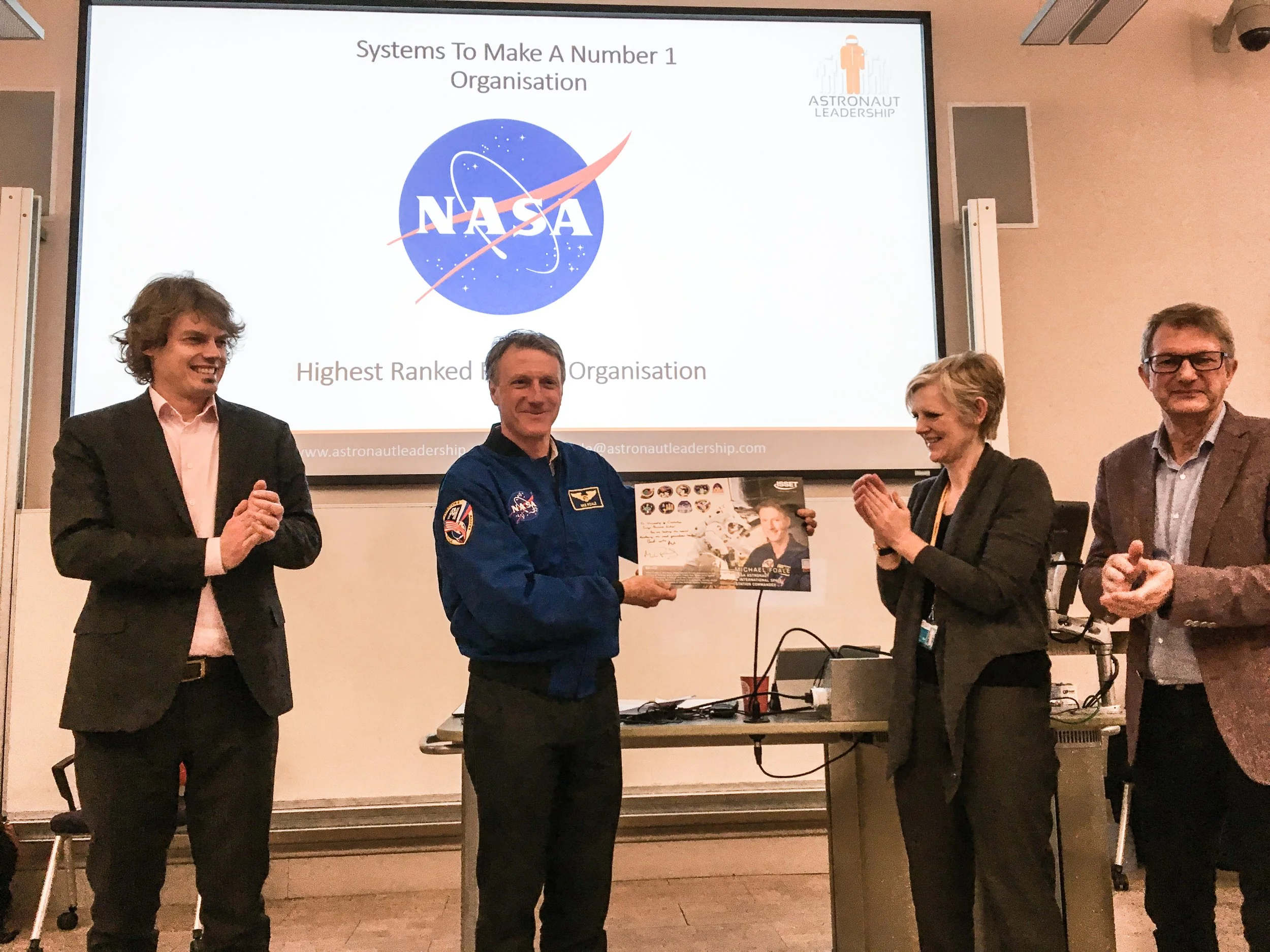 Group of four people standing in a classroom, with one person dressed in a NASA astronaut suit, holding a framed poster, and the others applauding. Behind them, a presentation slide displays the NASA logo and text about organization rankings.