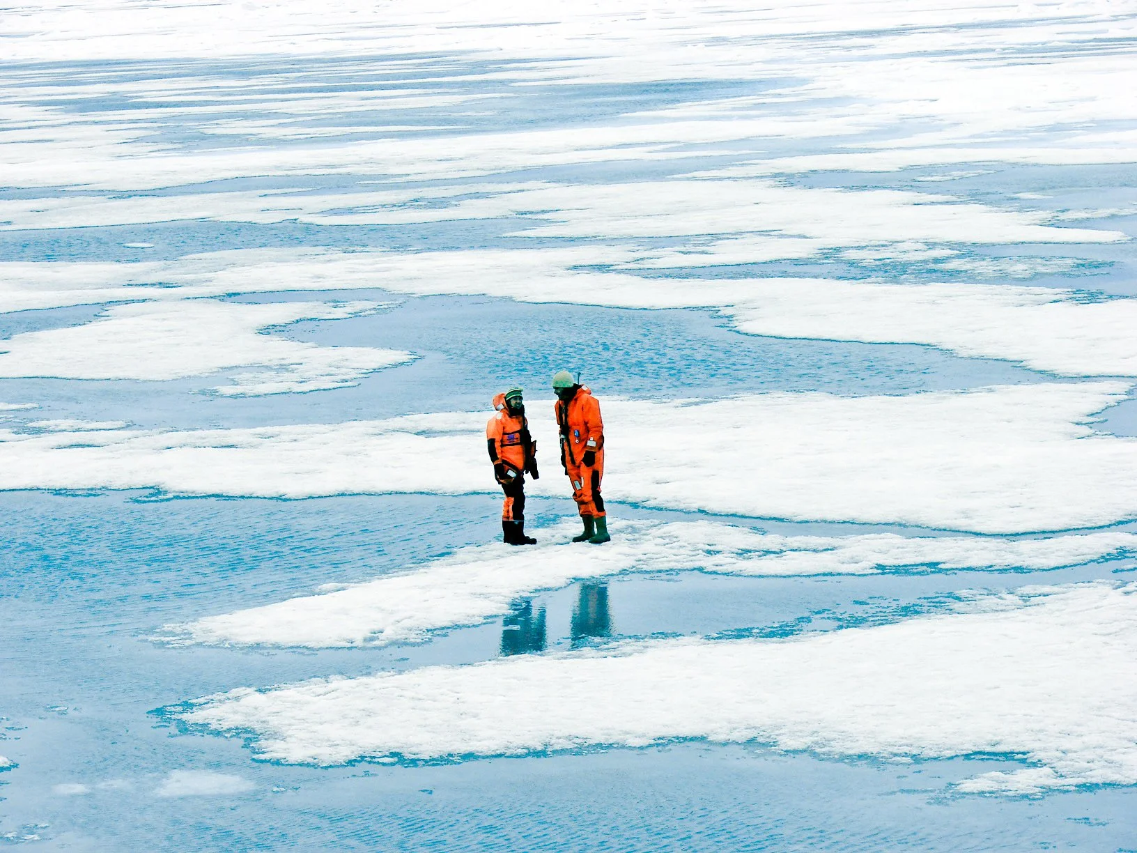 Two scientists in orange winter suits and helmets standing on a cold, icy landscape with floating ice patches on water.
