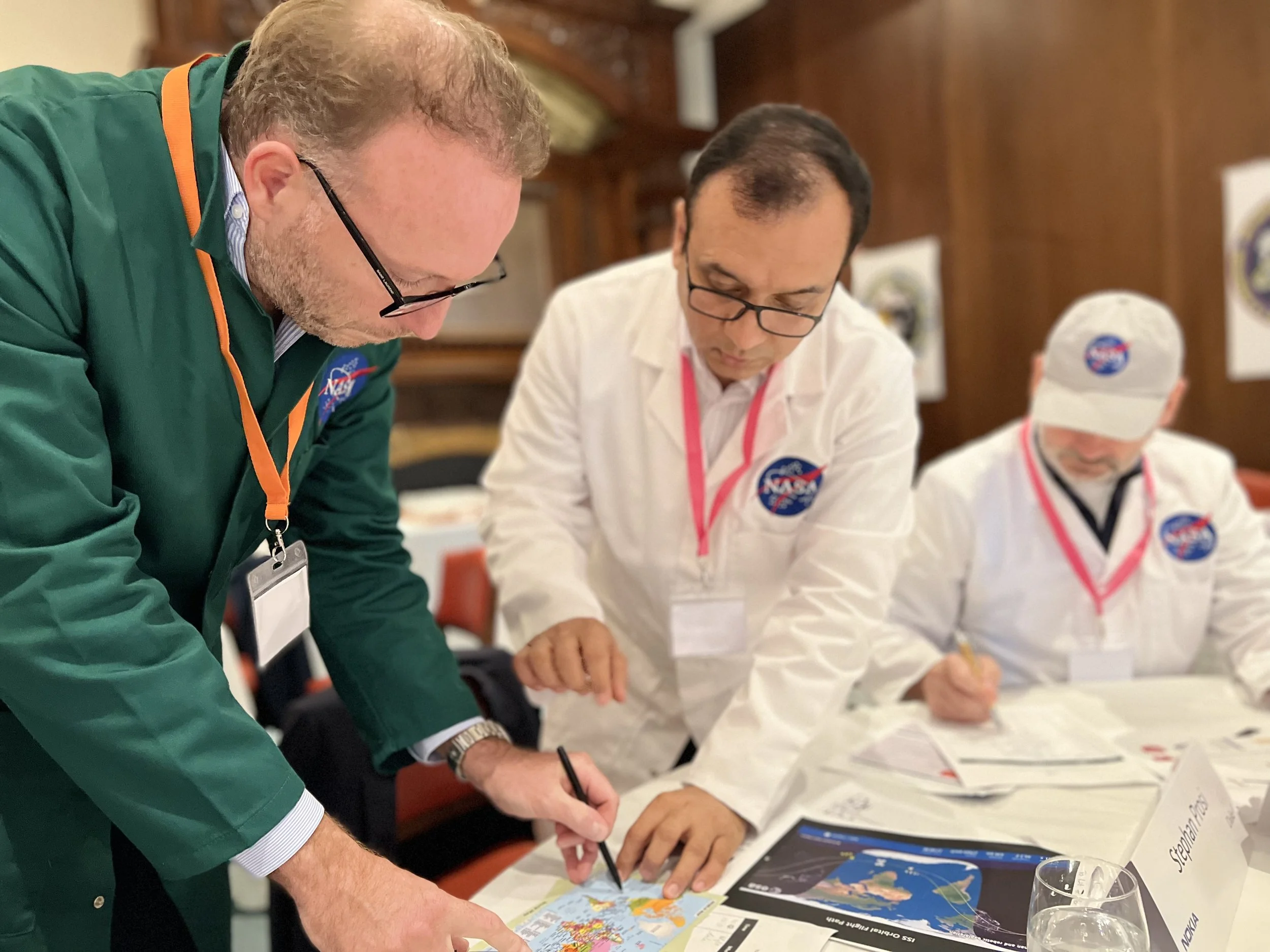 Three men in NASA outfits working on a map and weather chart on a table, in an indoor setting with wooden walls.