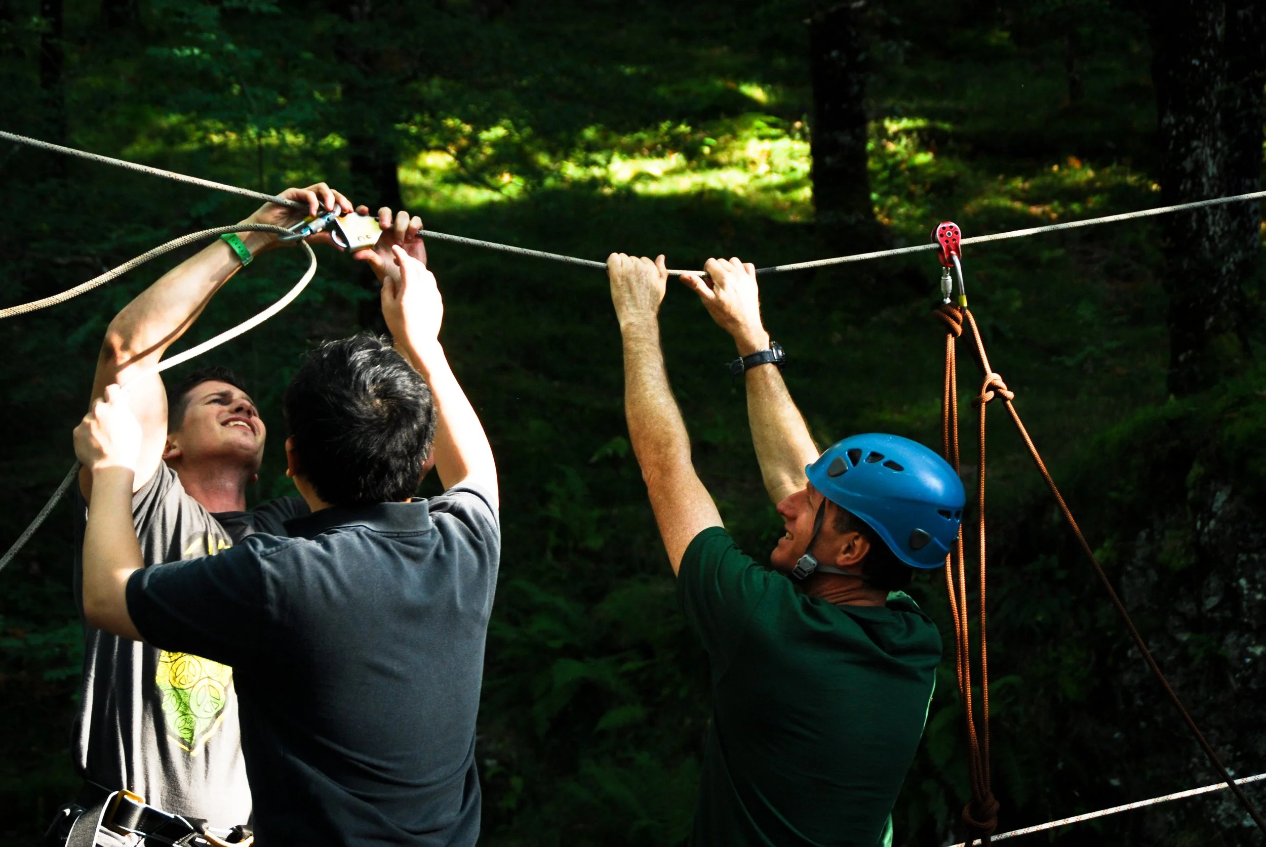 Three men in a forested area set up safety harnesses and ropes, with one wearing a blue helmet, for a zip-lining activity.