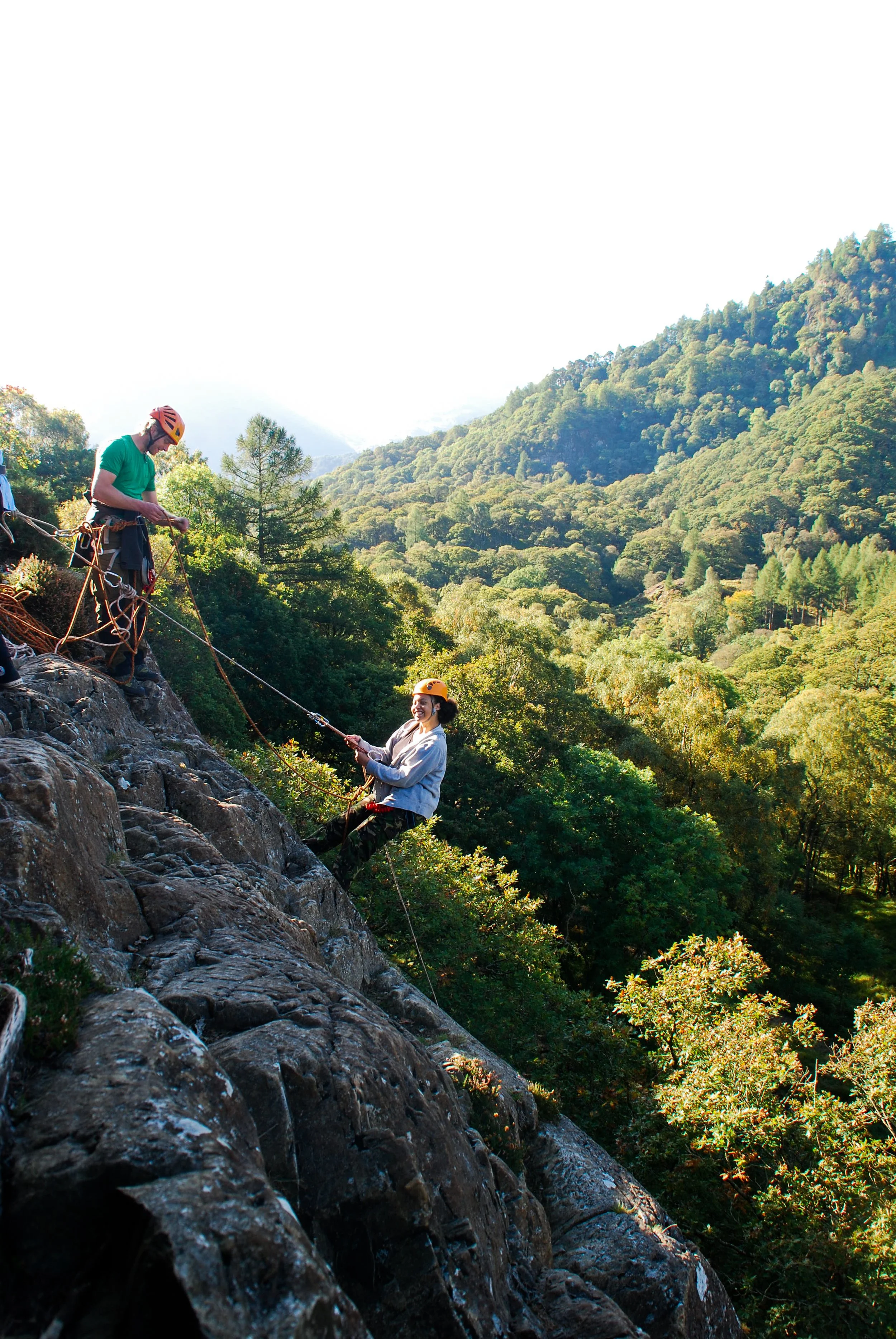 Two people rock climbing outdoors on a rocky slope, with a forested mountain range in the background; one climber is securing the ropes while the other is climbing.