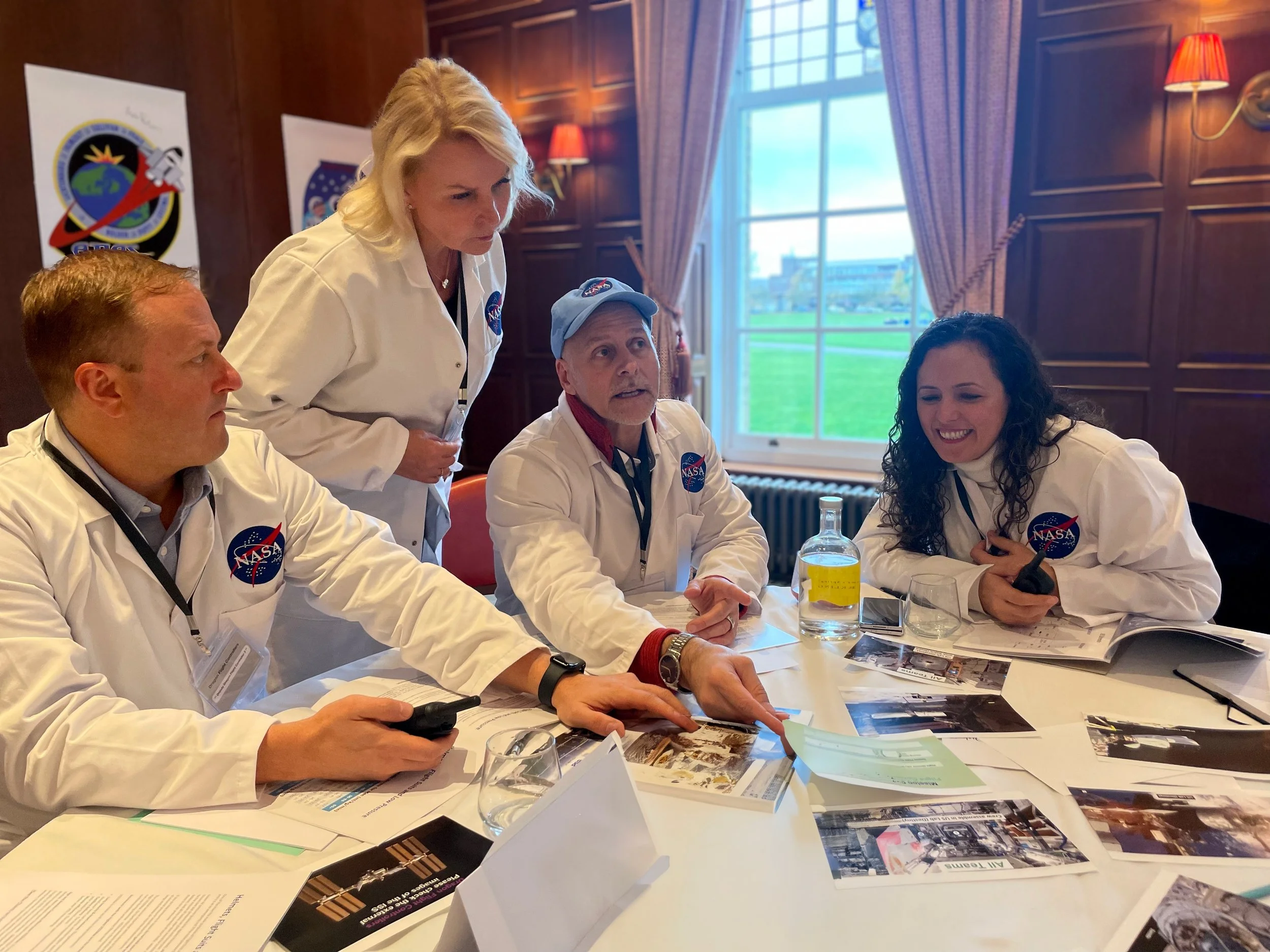 Four NASA staff members in white lab coats sitting at a table covered with documents and photographs, with one person standing and another smiling, in a wood-paneled room with a large window.
