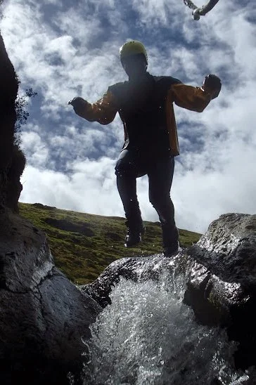 Person wearing yellow hat and dark outdoor clothing jumping over a small stream in a natural landscape with cloudy sky.