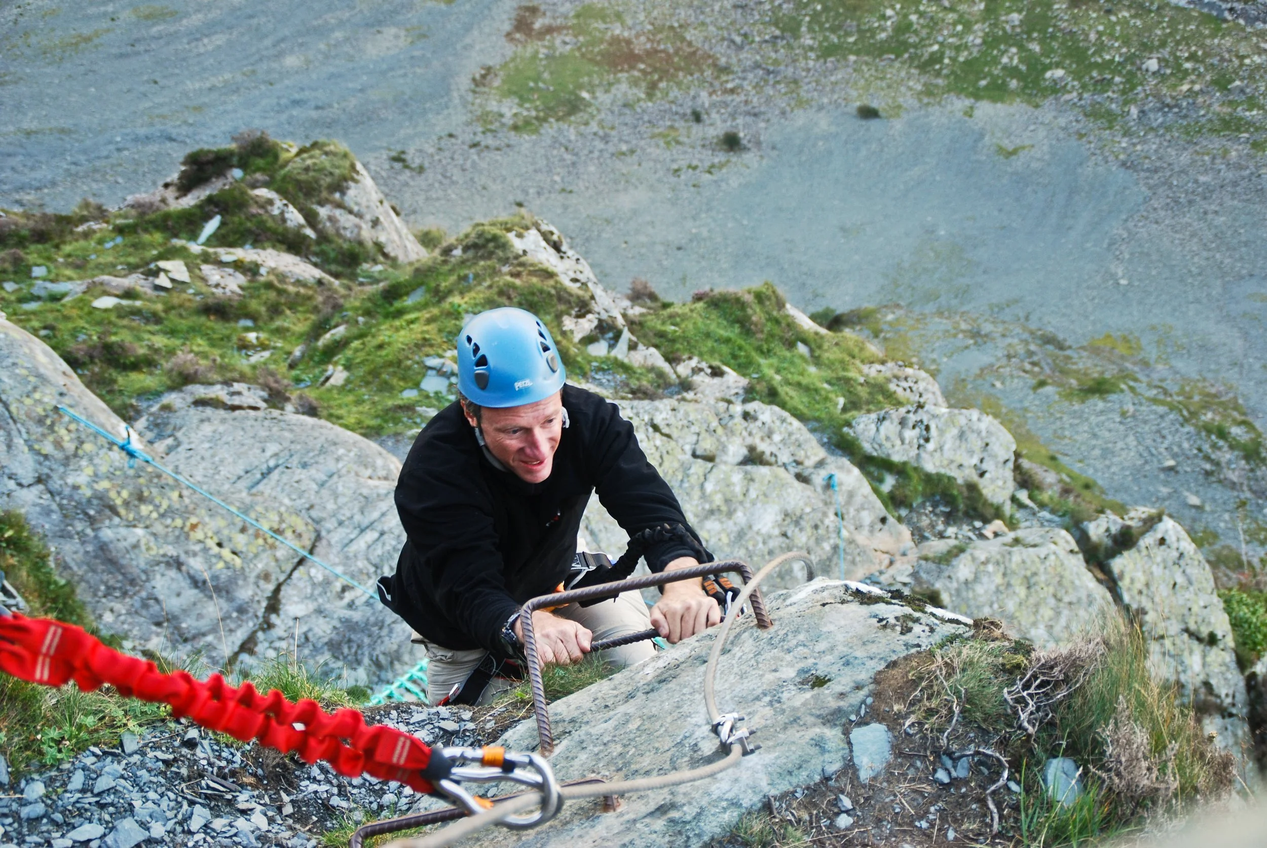 Man climbing a rocky outdoor mountain with safety equipment, wearing a blue helmet and black jacket.