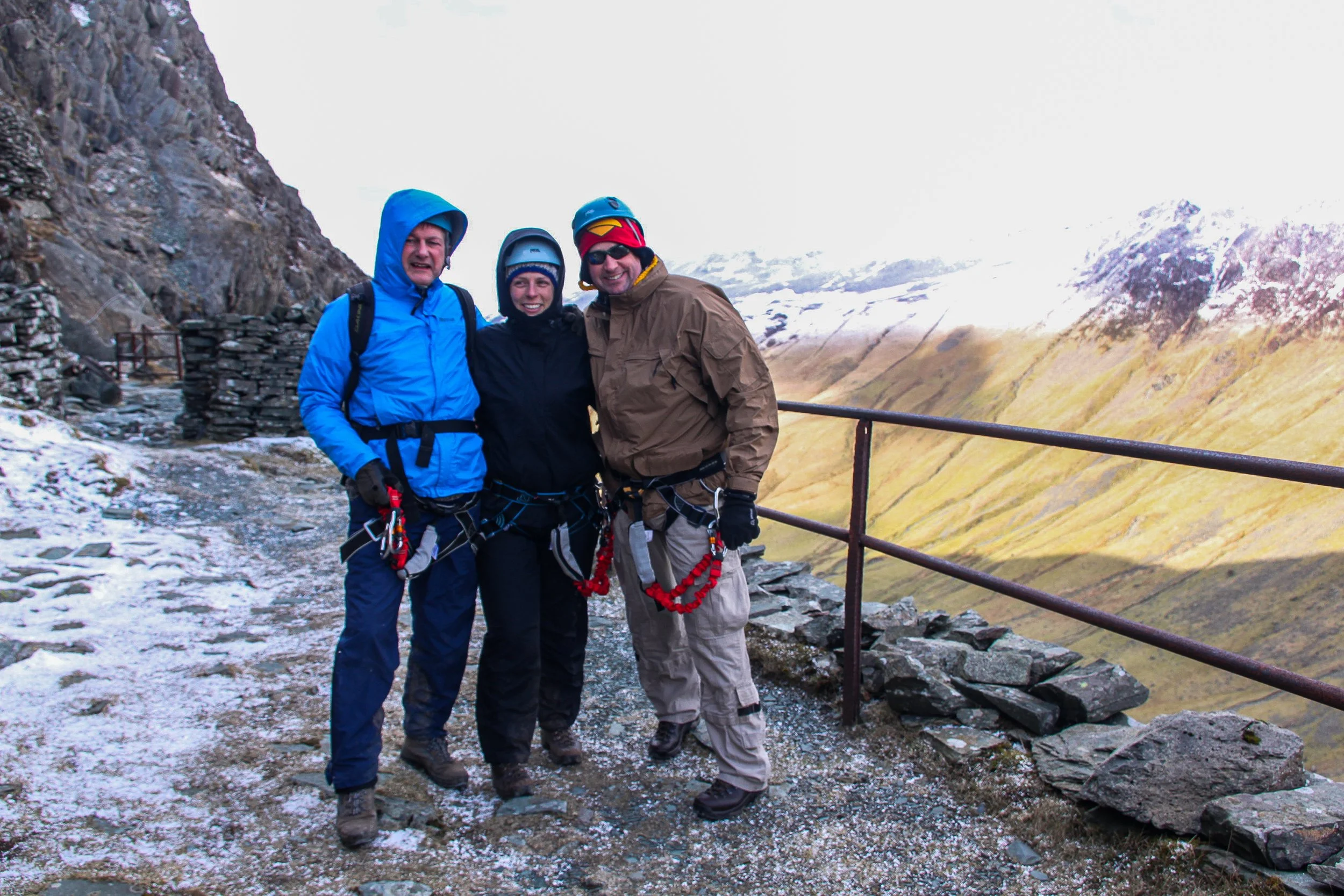 Three adventurers in outdoor gear stand together on a mountain trail with snow and rugged landscape in the background. They are smiling and wearing harnesses and helmets.