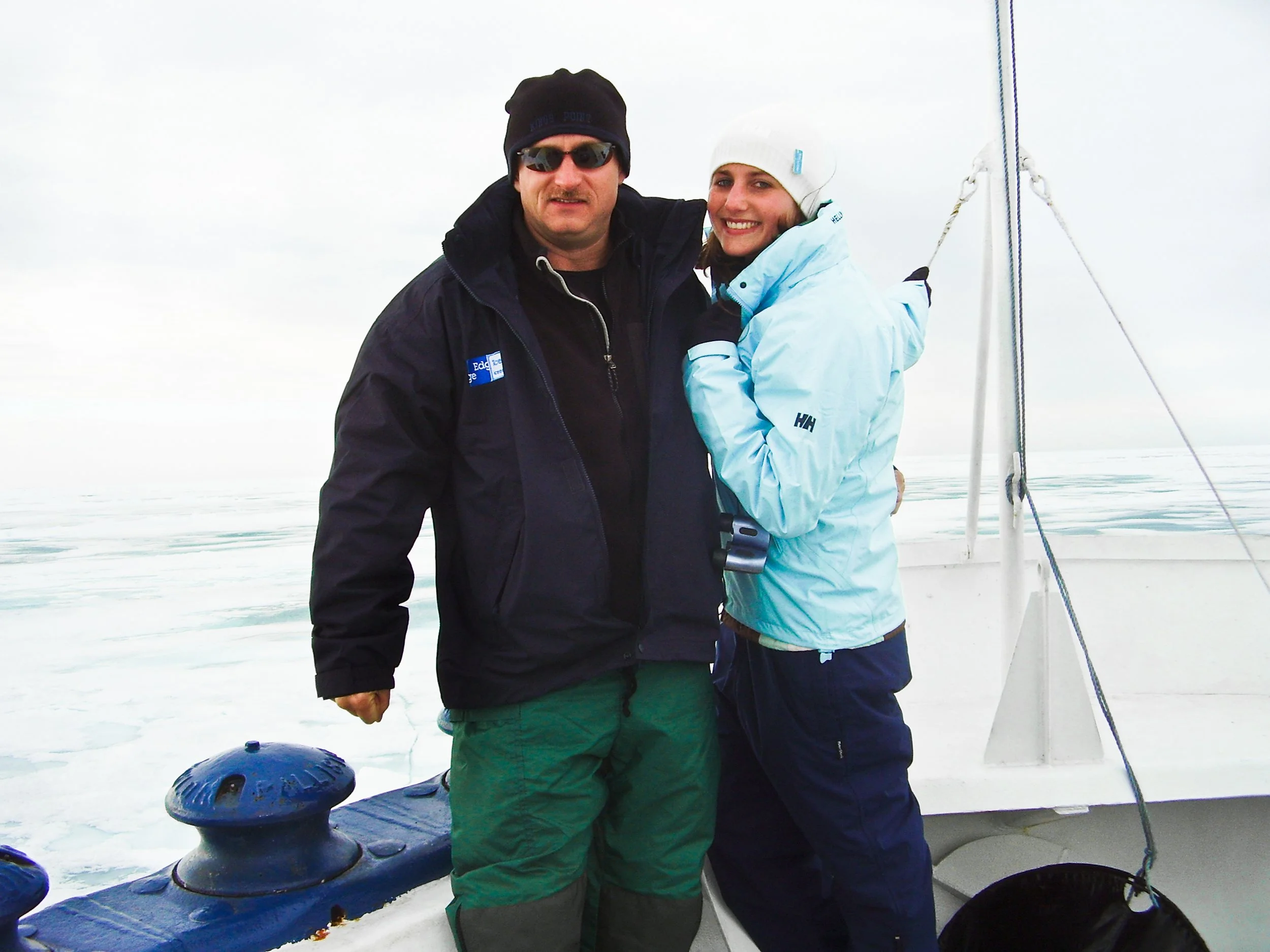 Two people standing on a boat in a cold, icy environment, smiling at the camera; one wearing a black jacket and sunglasses, the other in a light blue jacket and white hat.