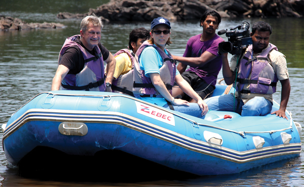 Group of six people in a blue inflatable raft, some wearing life jackets, on a body of water with rocks in the background, one person holding a video camera.