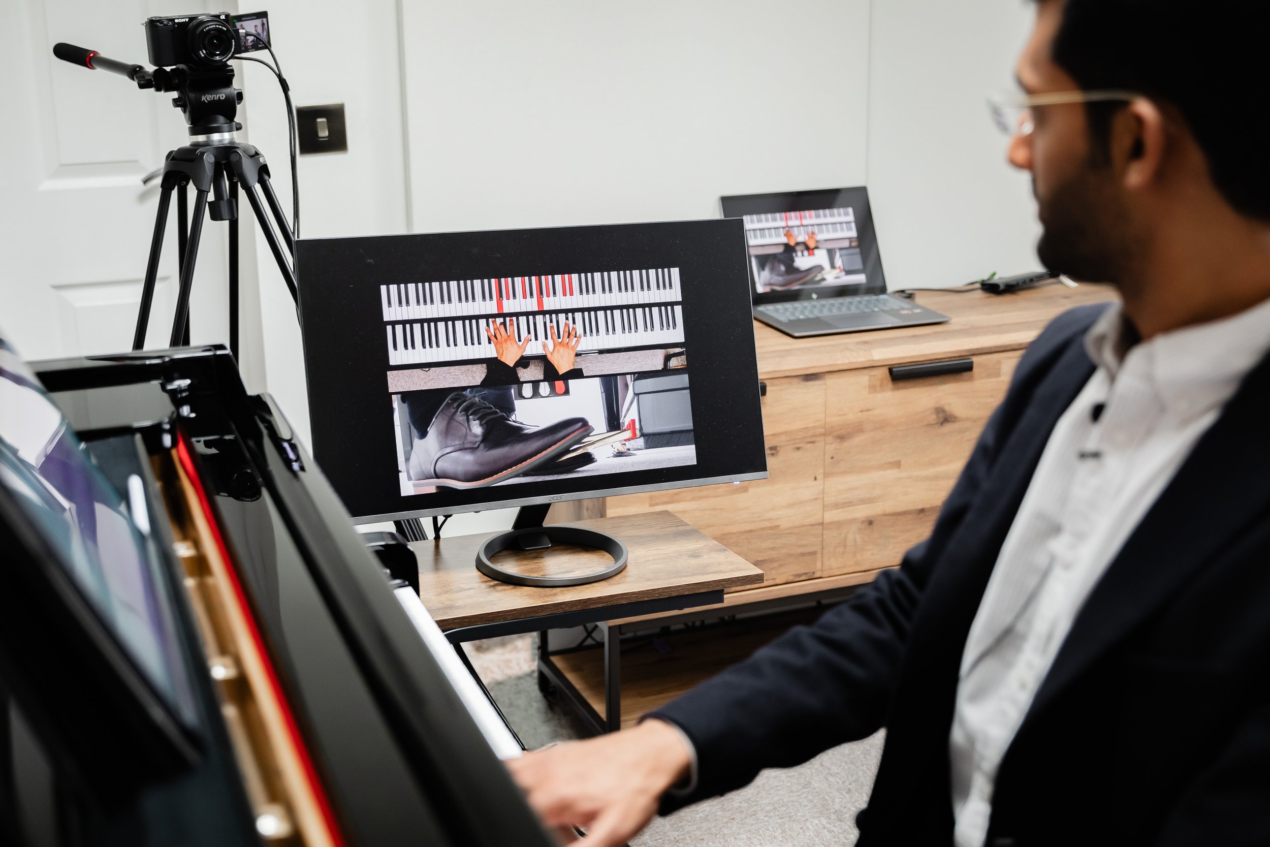 A Virtuoso Project piano tutor playing digital piano with a computer screen showing a virtual piano tutorial, a camera mounted on a tripod capturing the scene, and a laptop in the background.