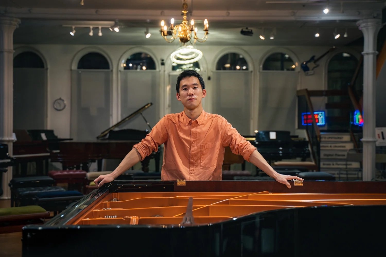 A young man in an orange shirt standing behind an open grand piano in a music room, with other pianos and music equipment in the background.