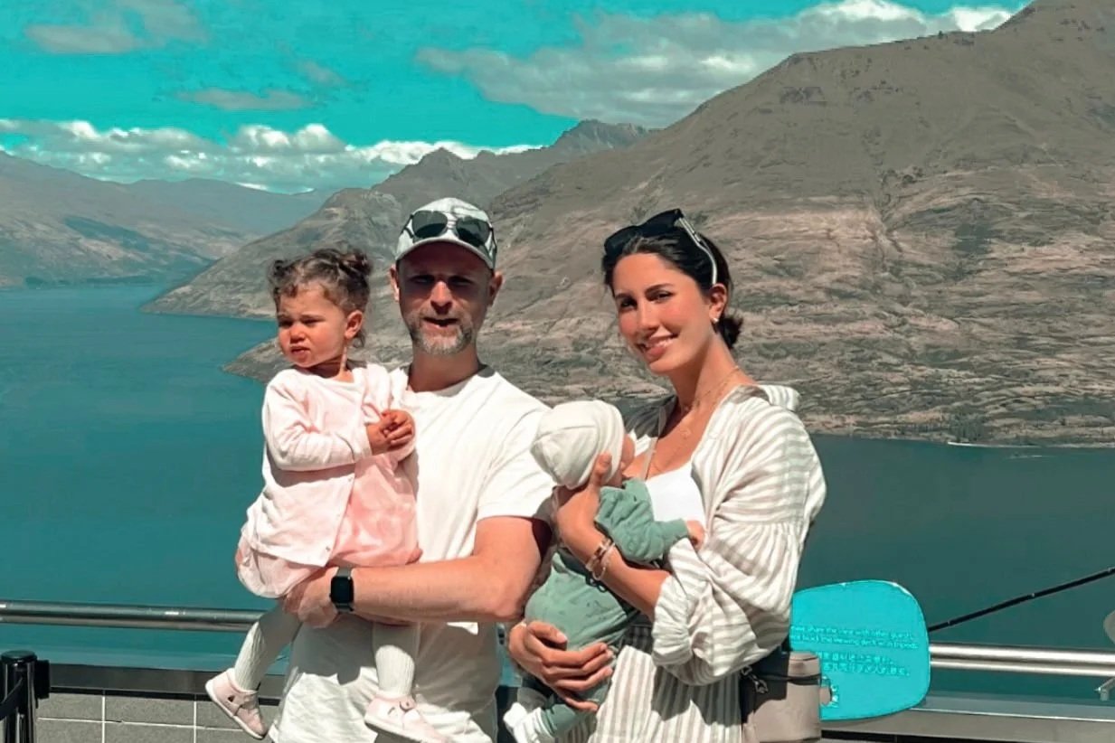 A family of four standing on a viewing platform with a lake and mountains in the background. The father is holding a young girl, and the mother is holding a baby. The sky is partly cloudy.