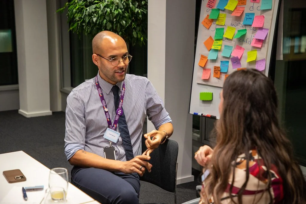 A man in a business shirt and tie, wearing glasses, sitting at a table engaged in conversation with a woman with long hair in a patterned top. They are in an office or meeting room with a whiteboard covered in colorful sticky notes behind them, and a window with a plant outside.