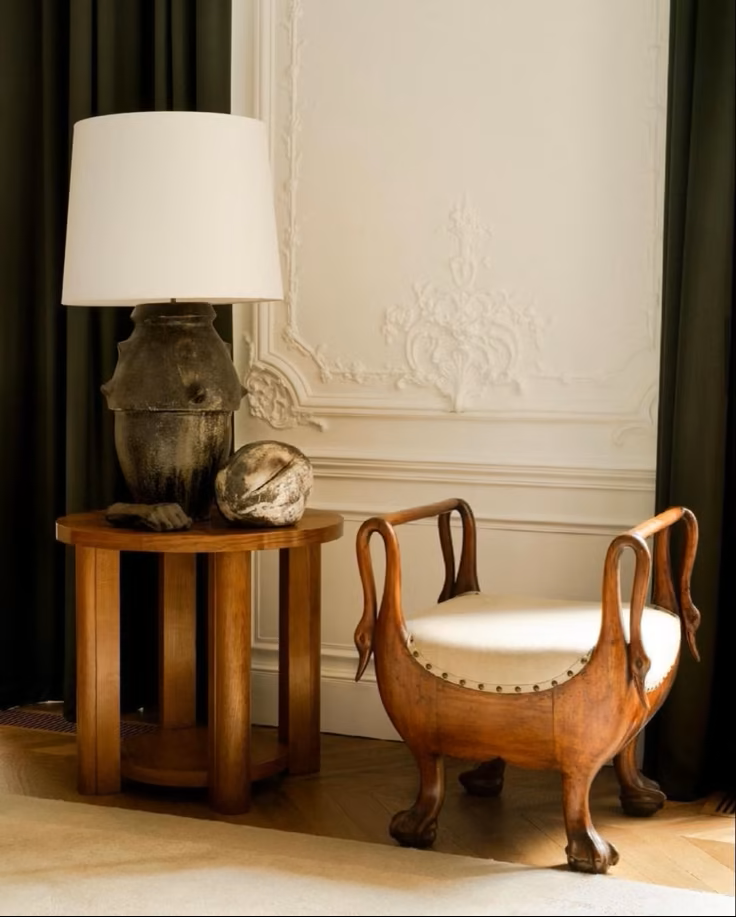 A wooden round side table with an amphora lamp with a white shade, placed next to a beautiful antique french scroll carved wooden stool with a white cushioned seat in an elegant room with ornate wall details.