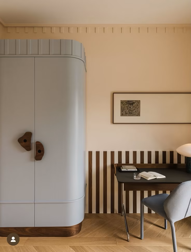 A minimalist interior corner featuring a white curved wardrobe with wooden handles, a striped wooden paneling on the wall, a small black desk with items, an open book, a glass of water, and a gray upholstered chair.
