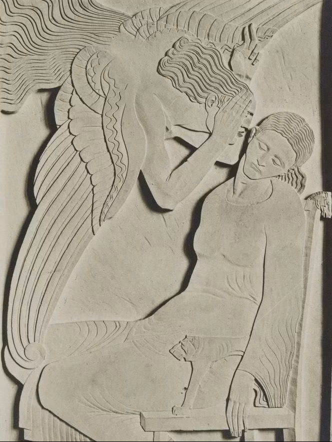 A carved stone relief depicting a woman sitting on a bench with a serene expression, and an angelic figure leaning over her, touching her forehead. The scene has detailed carved lines showing flowing hair and draped clothing.