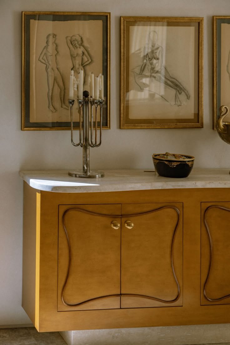 A wooden sideboard with a marble top, decorated with a candle holder and a bowl, hanging framed sketches of women on the wall behind it.