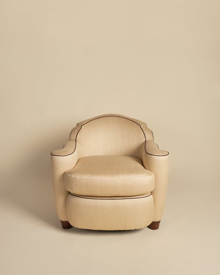 A beige upholstered armchair with dark piping on a beige background.
