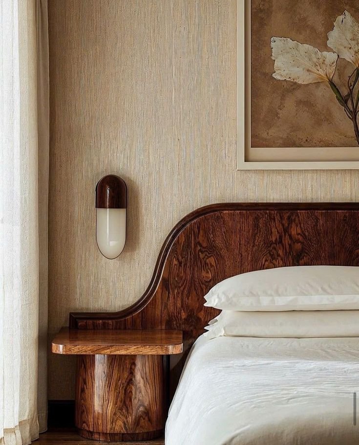 Close-up of a bedroom corner featuring a wooden headboard, white bedding, a wall-mounted light, and a framed botanical print.
