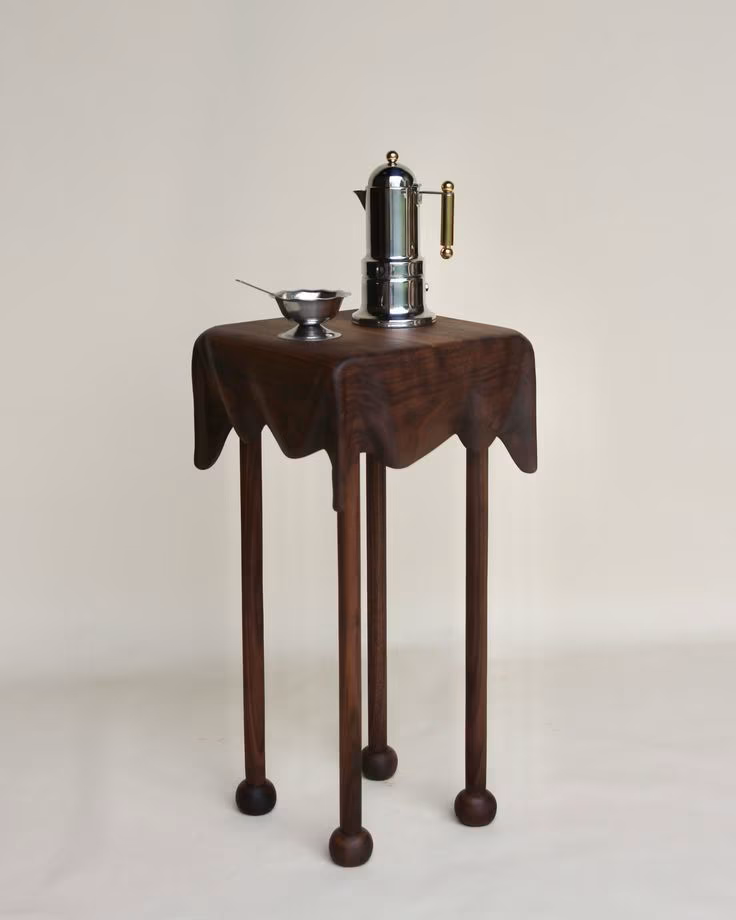 A wooden table with a dark finish, holding a vintage coffee grinder and a small metal bowl with a spoon, against a plain light background.