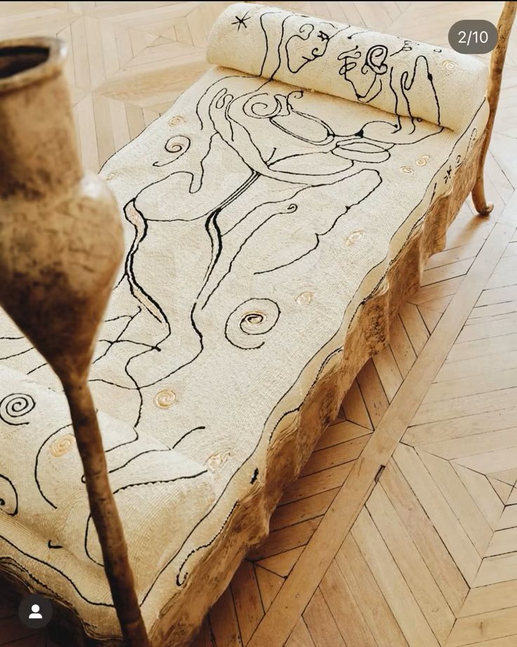 An antique decorative day bed with a beige fabric cover featuring black line art of a woman holding a cup, surrounded by swirling patterns, and a wooden frame with a natural, rustic finish.