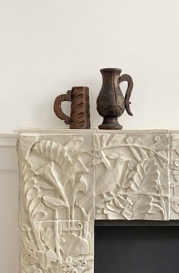 Decorative plaster relief fireplace mantel with carved floral and leaf patterns, topped with two primitive wooden vases, one brown and one dark brown.
